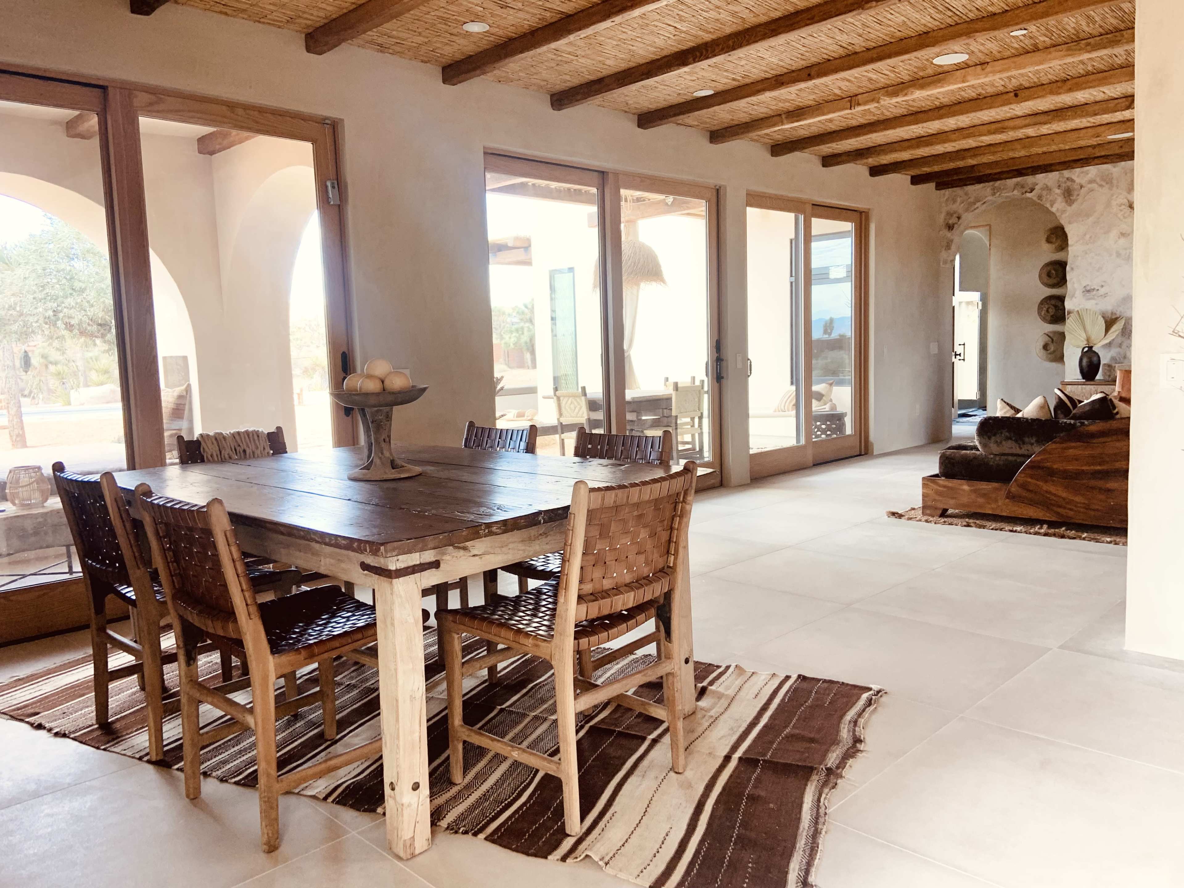 The interior features a wooden dining table with woven chairs, surrounded by large windows that let in natural light, and a spacious living area with a dark sofa and decorative elements.