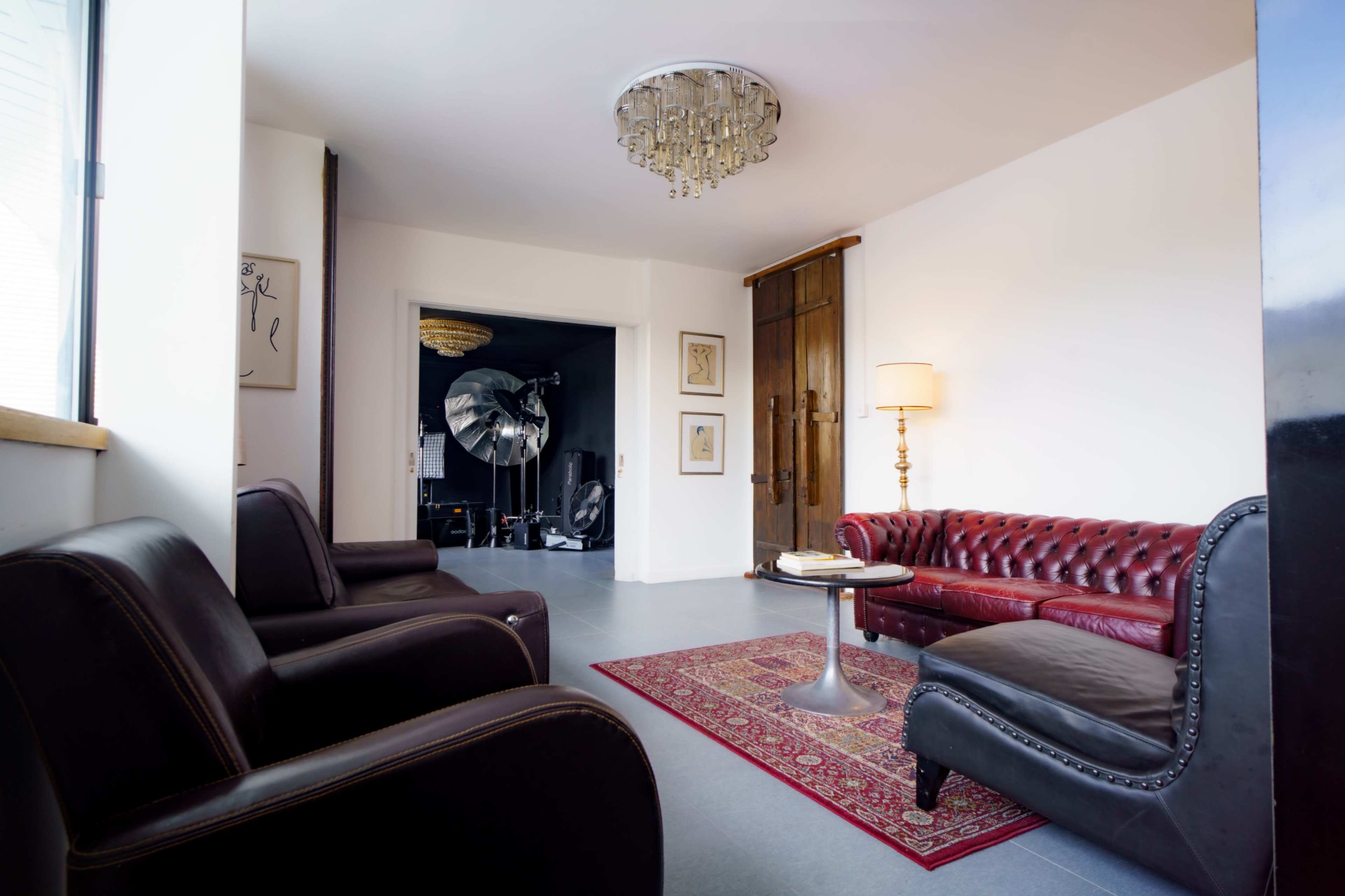 The image shows a modern interior of a living space featuring a red leather chesterfield sofa, a round coffee table on a patterned rug, and two dark leather armchairs, with a decorative chandelier overhead.