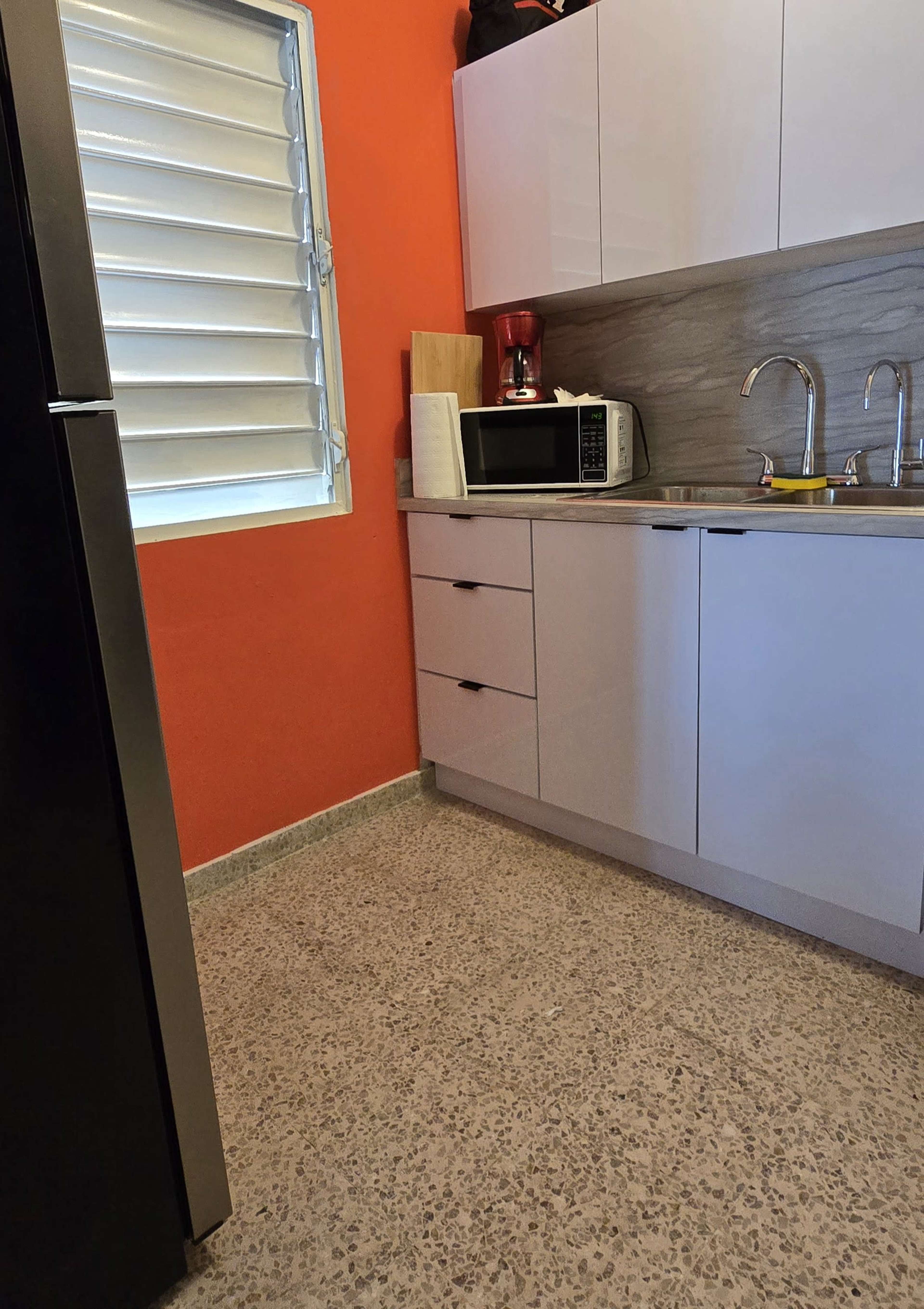 The image shows a small kitchen area with a refrigerator, microwave, sink, and cabinets, set against an orange wall.