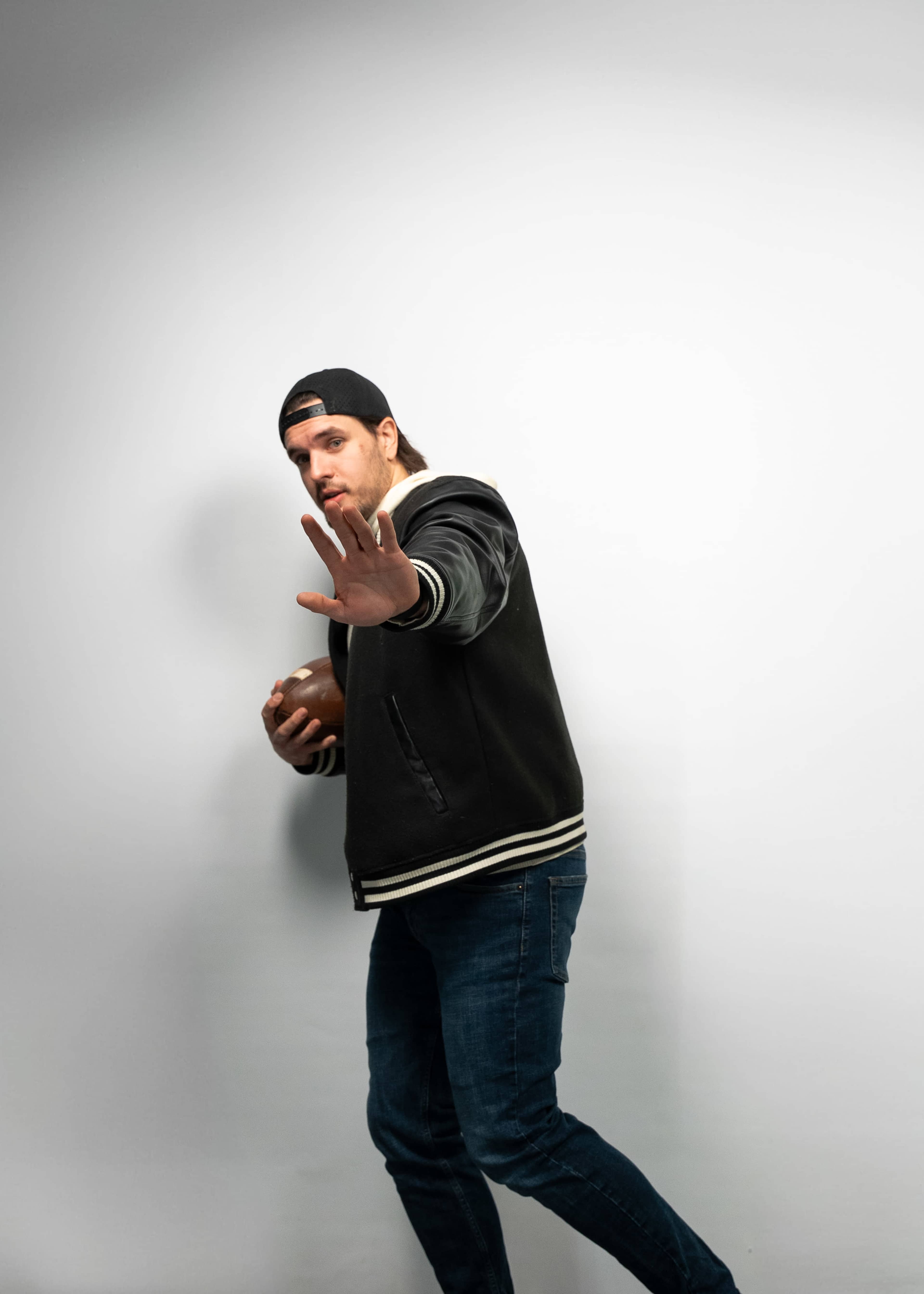 A man in a black varsity jacket and baseball cap stands against a plain white background, holding a football and raising his hand in a stopping gesture.