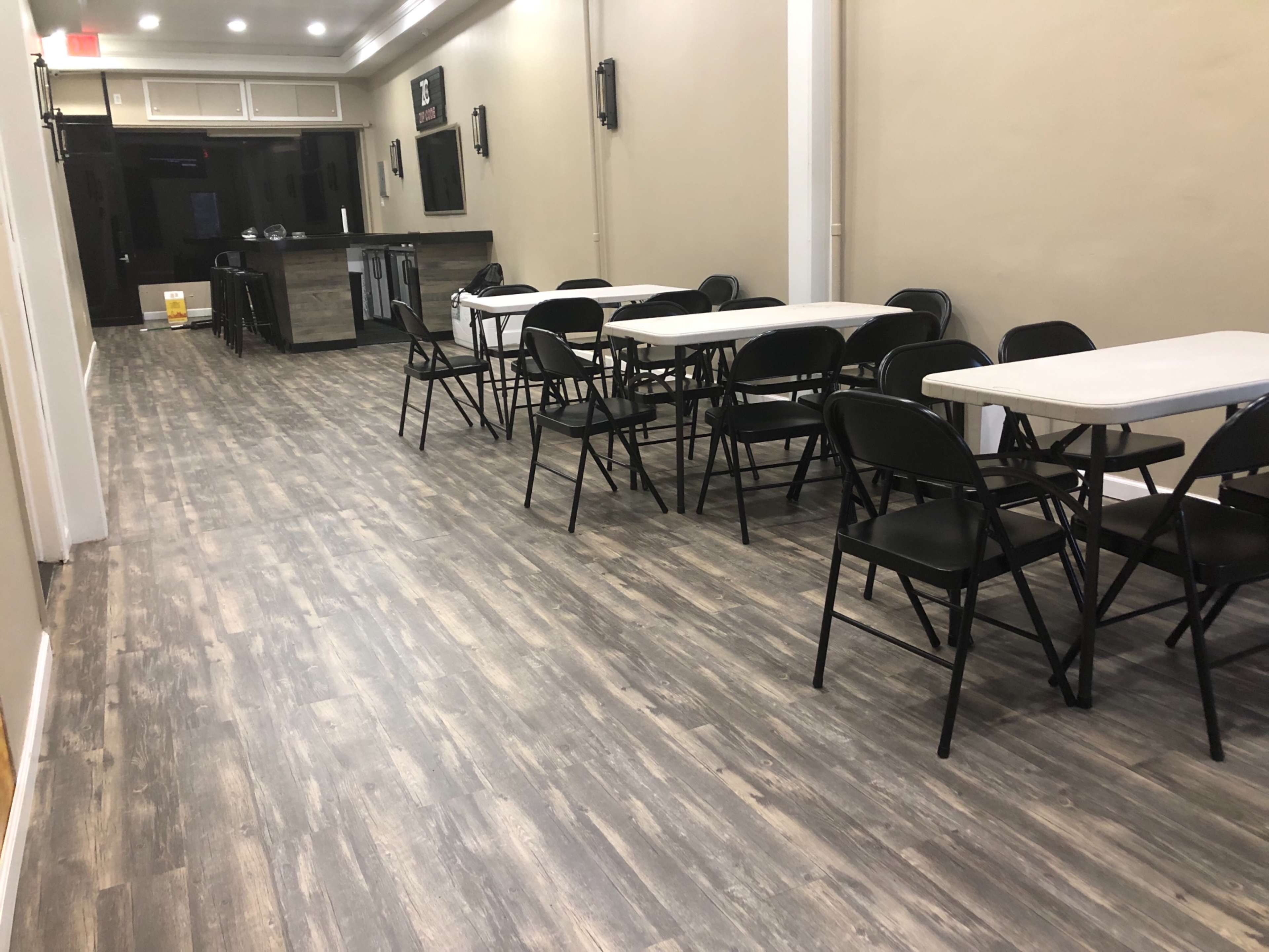 The image shows a narrow room with wooden flooring, featuring several tables and black folding chairs arranged in rows, leading toward a service area at the back.