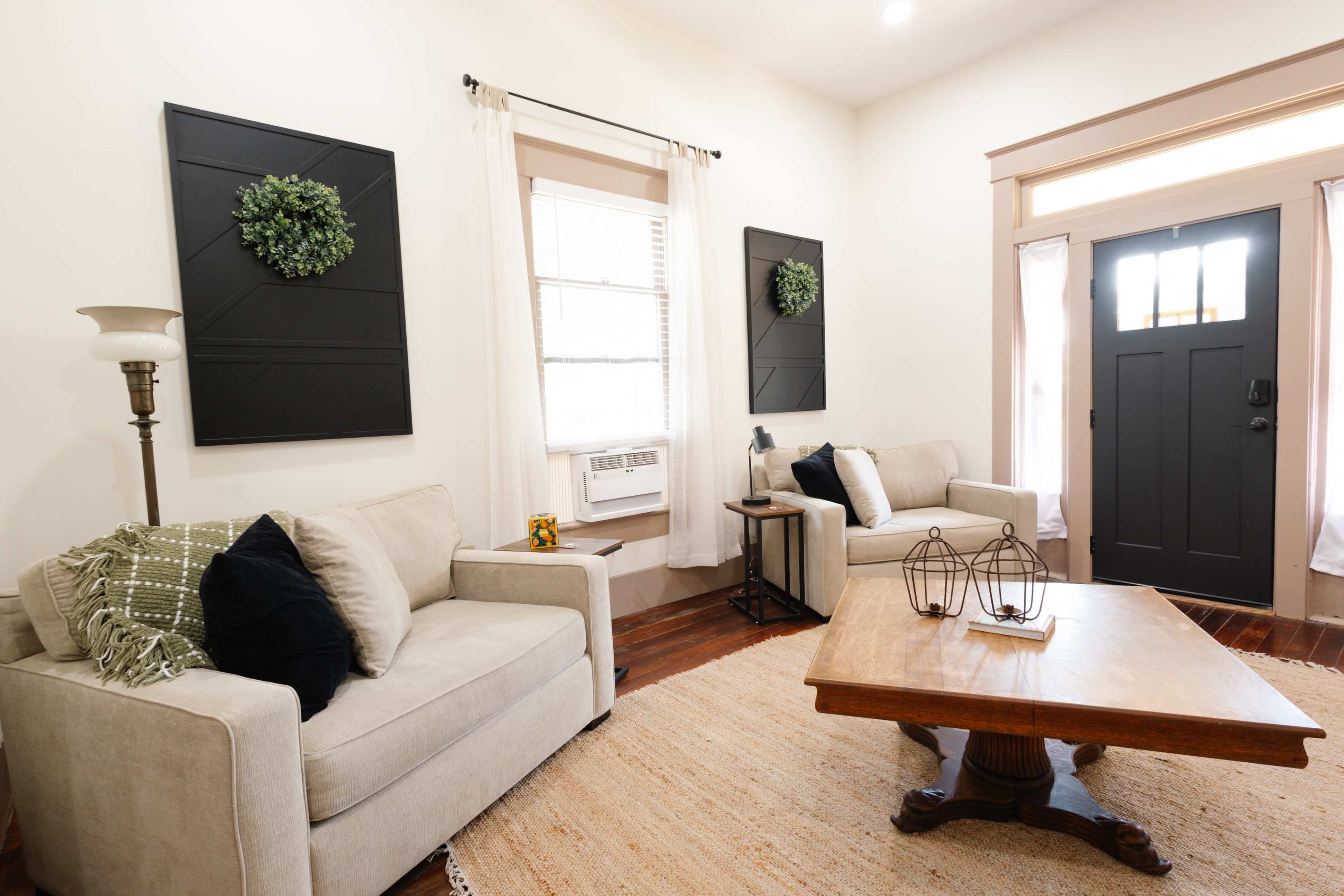 The image shows a cozy living room featuring two light-colored sofas, a wooden coffee table, an air conditioning unit, and decorative wall art, with natural light streaming through a door.