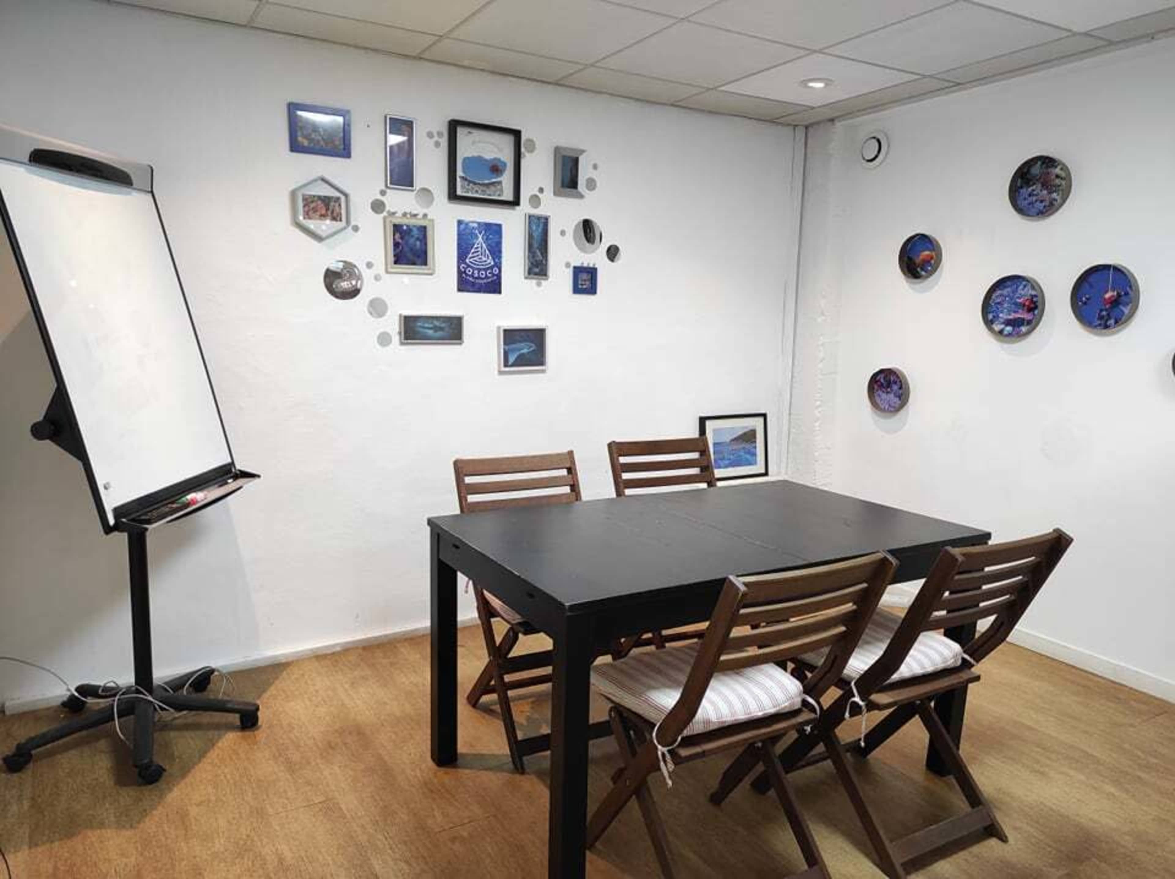 A meeting room features a black table surrounded by wooden chairs, with a whiteboard and framed pictures on the walls.
