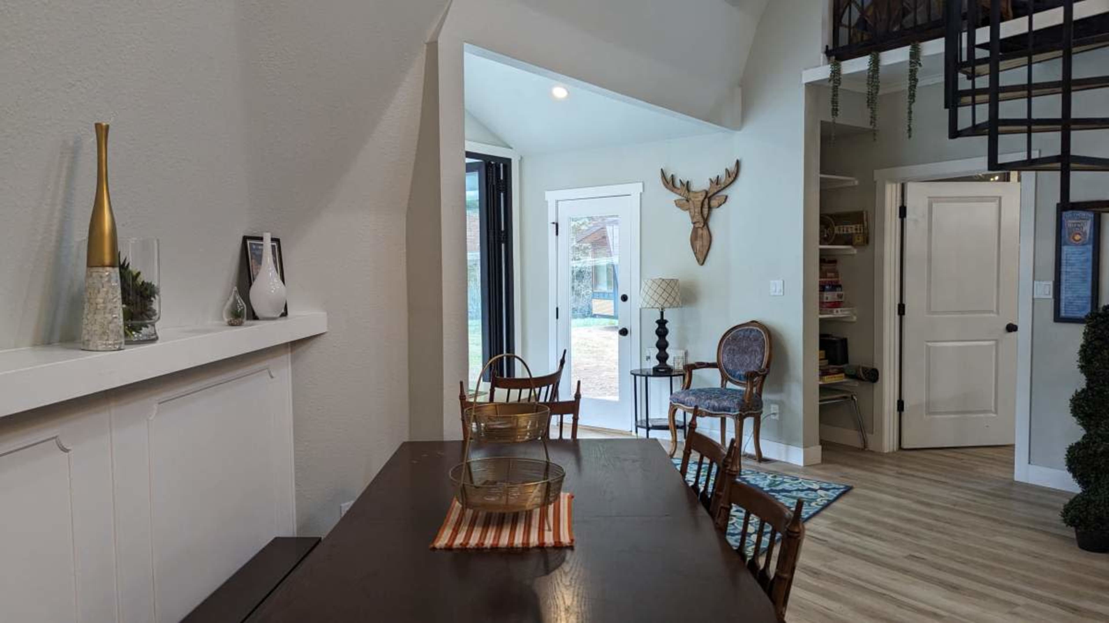 A dining area with a wooden table and chairs leads to a doorway that opens to the outdoors, featuring a wall-mounted deer head and a bookshelf.