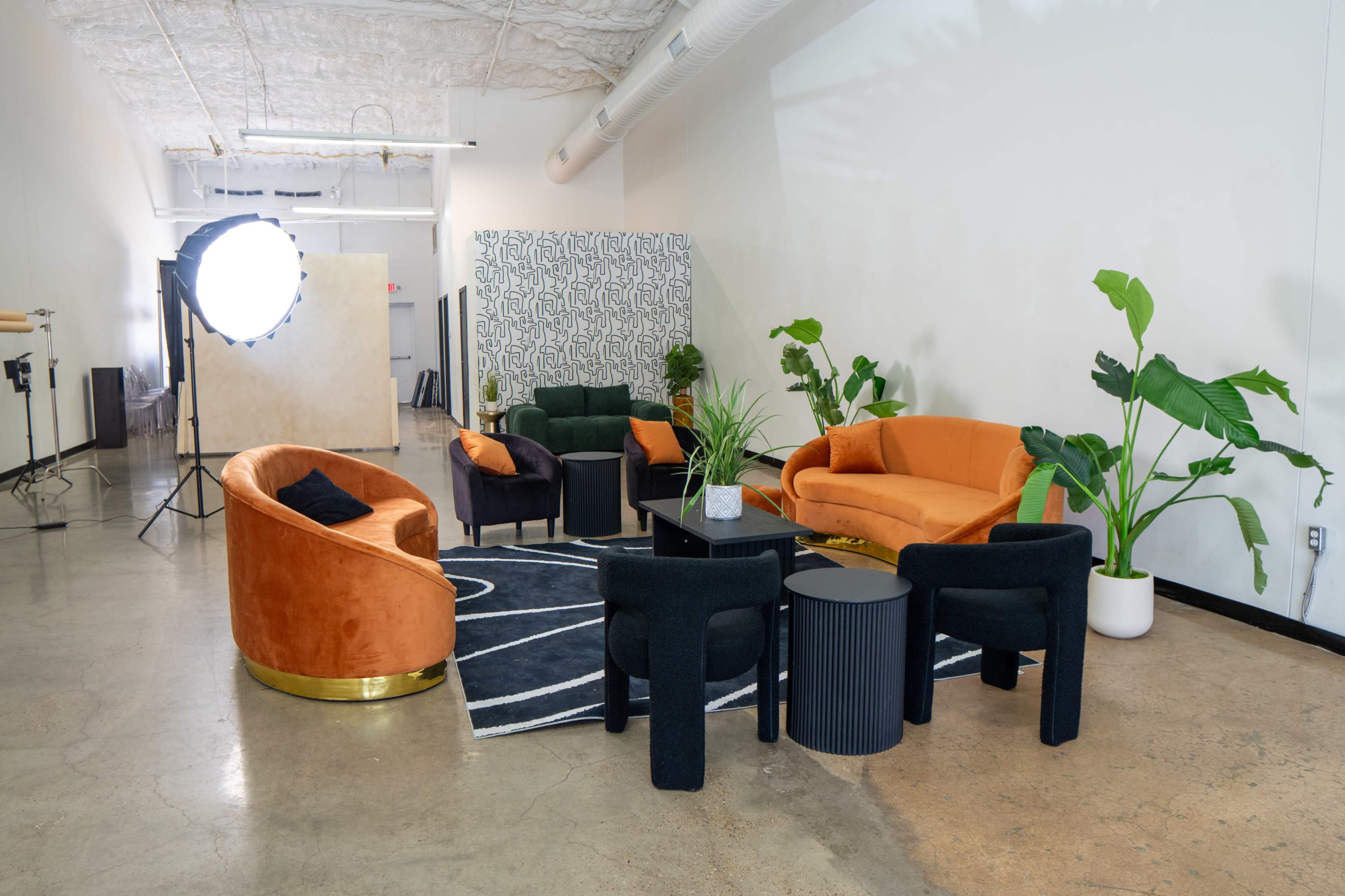 The image shows a modern lounge area with a combination of orange, green, and black seating arranged around a central table, surrounded by plants and a patterned backdrop.