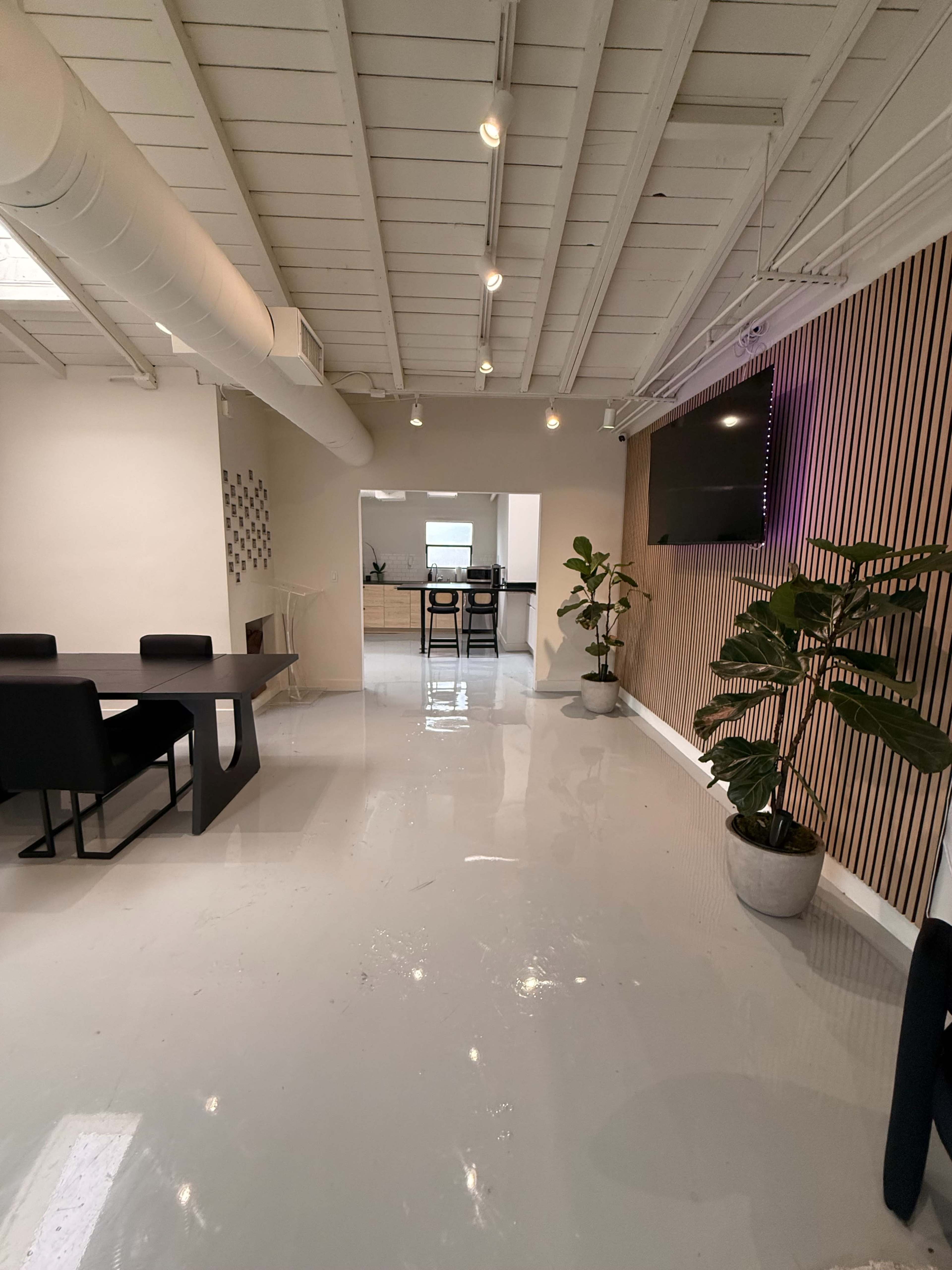 The image shows a modern office space with a glossy white floor, a black dining table, and a plant beside a wall featuring wood paneling and a mounted television.