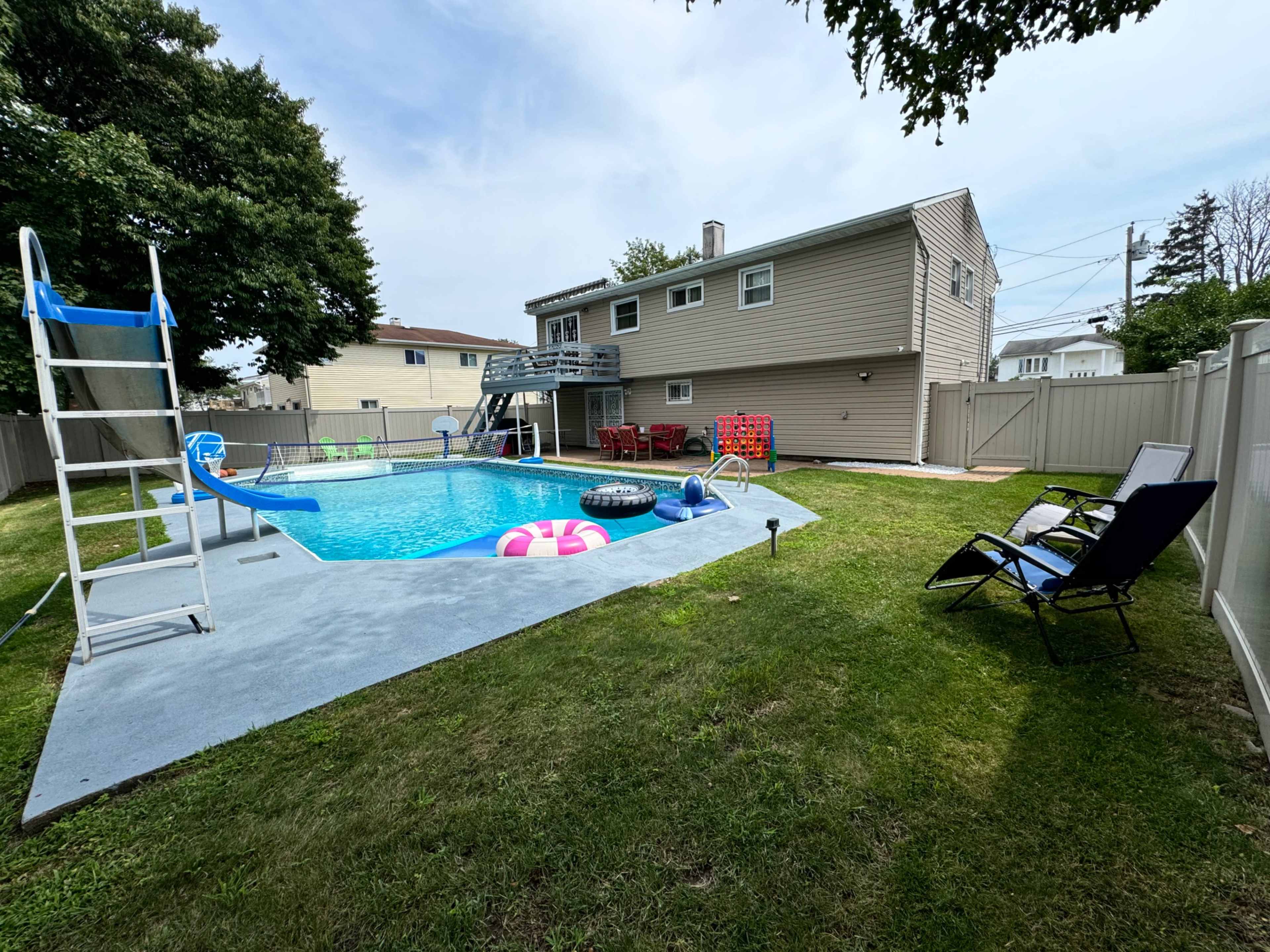 A backyard with a swimming pool, lounge chairs, and a small slide, surrounded by a fence and a two-story house in the background.