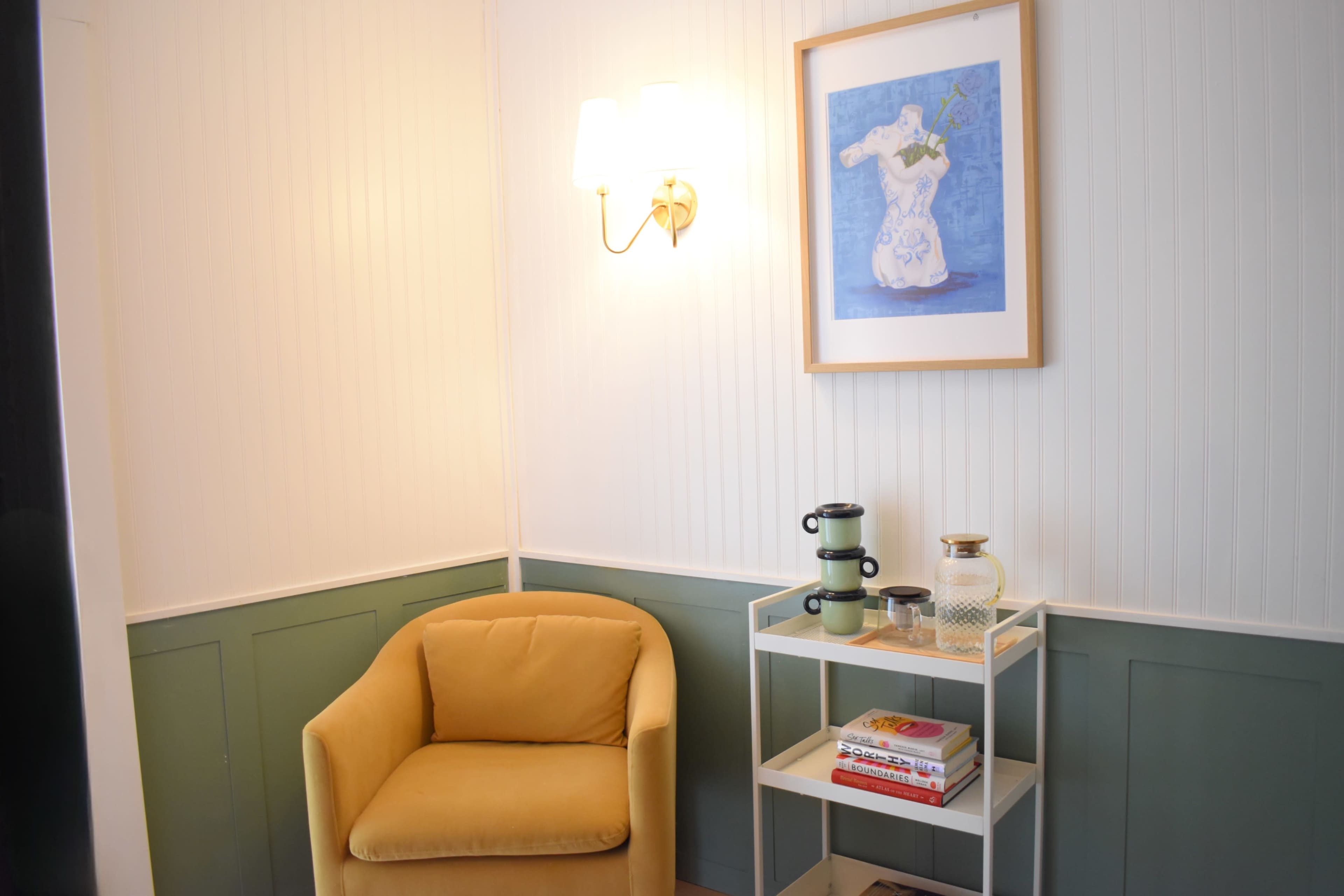A small corner of a room features a yellow armchair, a shelf with books and decorative items, and a framed artwork hanging on the wall.