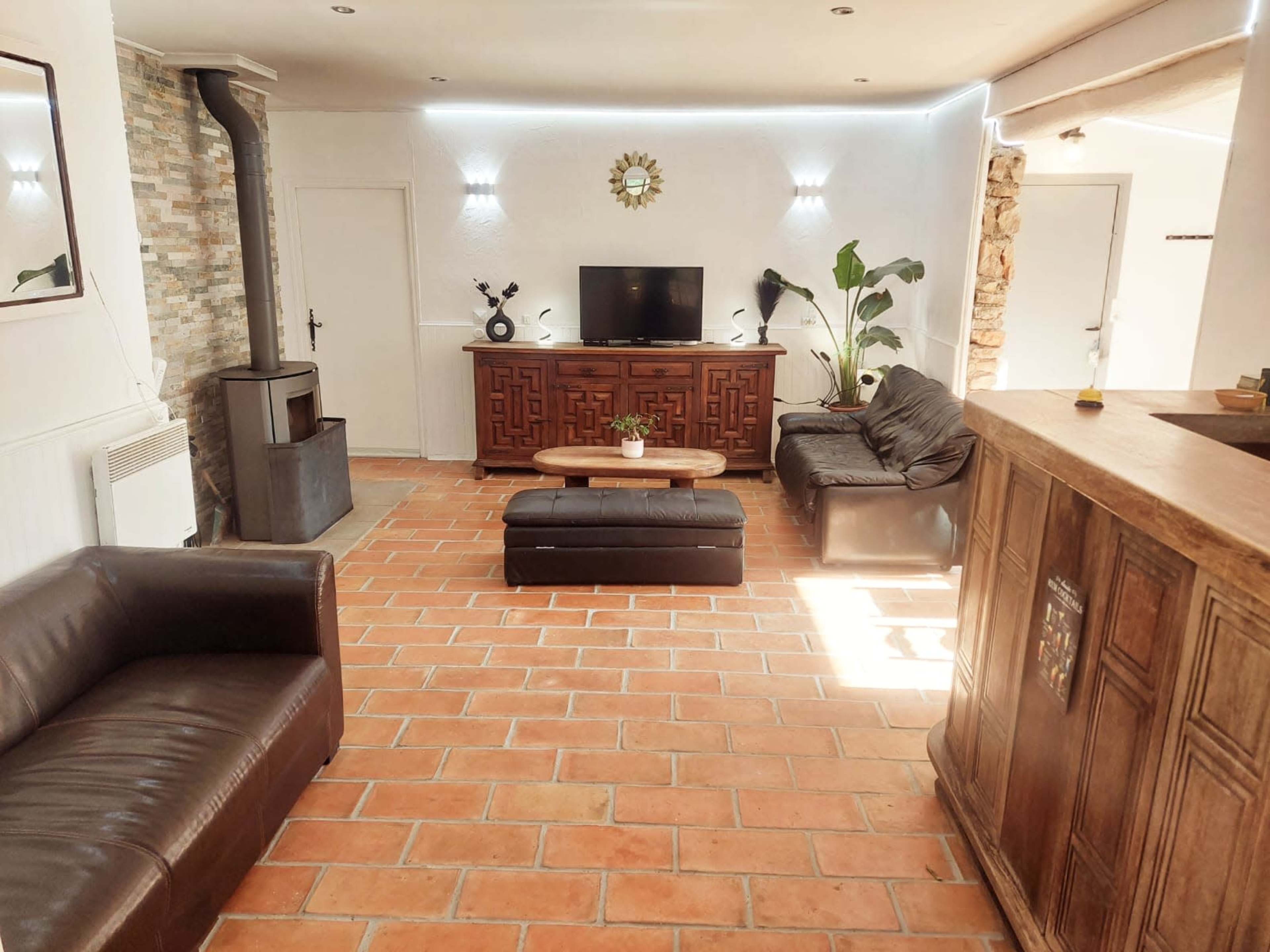 The room features a living area with brown leather sofas, a wooden cabinet, a television, and a wood-burning stove, all set against terracotta tiled flooring.
