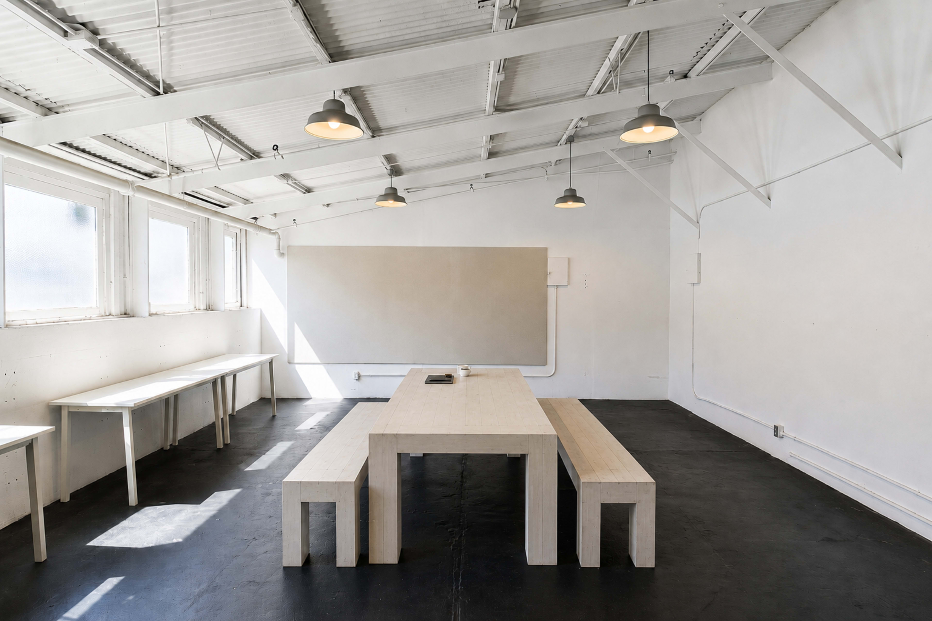 The image shows a spacious, minimalist room with a large wooden table and benches, illuminated by overhead lights and featuring white walls and a concrete floor.
