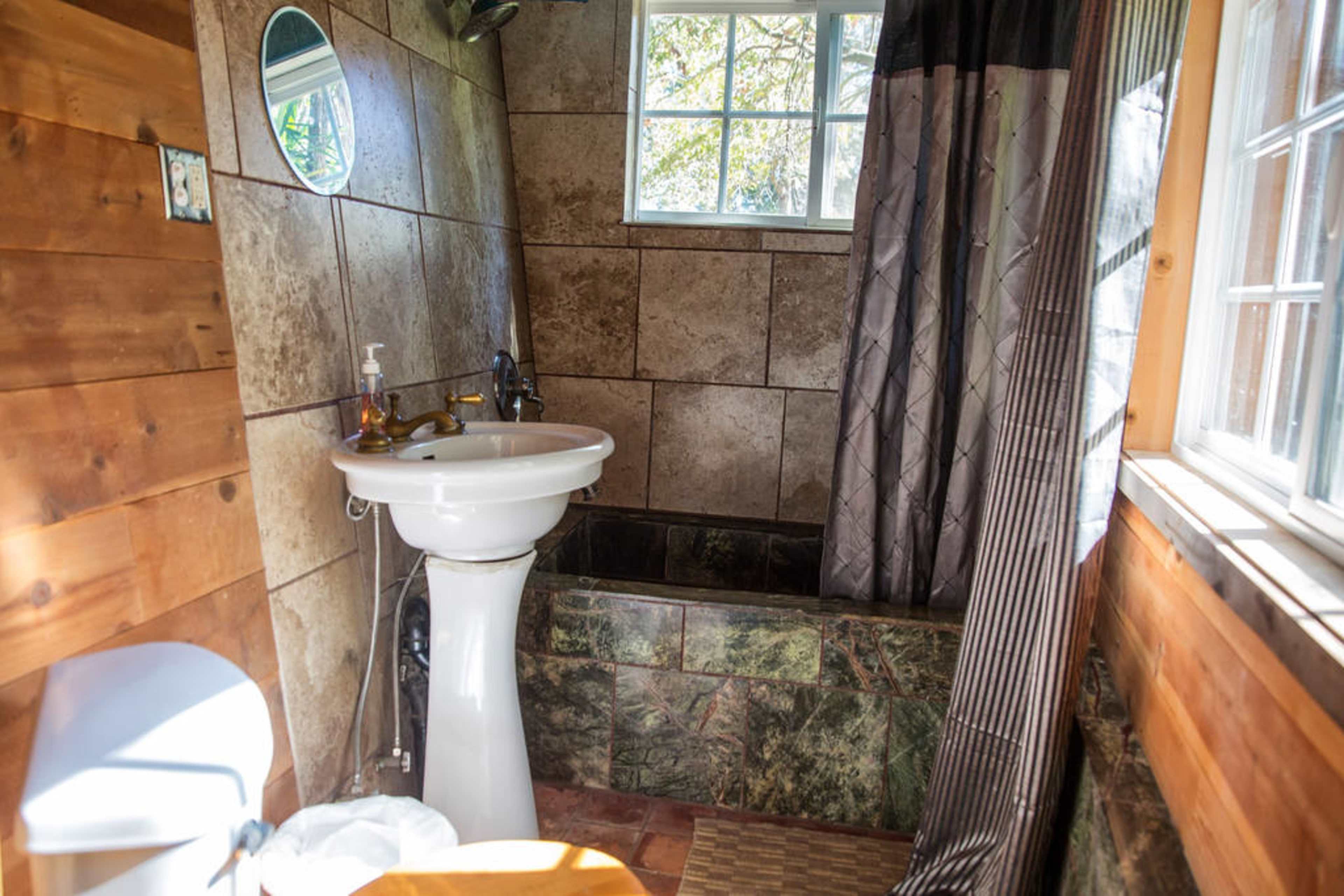 A bathroom with a pedestal sink, a shower area lined with stone tiles, and a window allowing natural light.