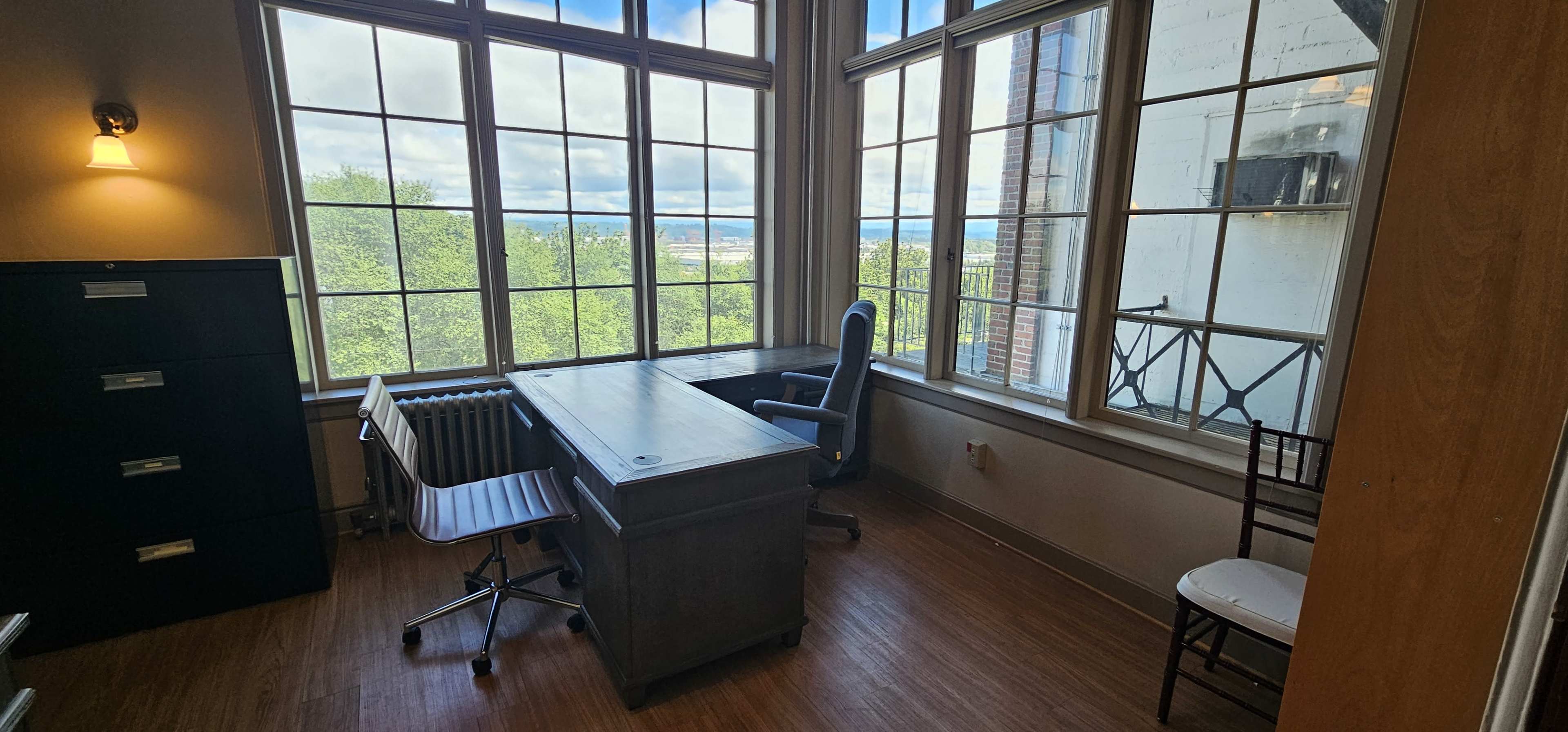 The image shows a bright office space with large windows, a wooden desk and a rolling chair, and a view of greenery outside.