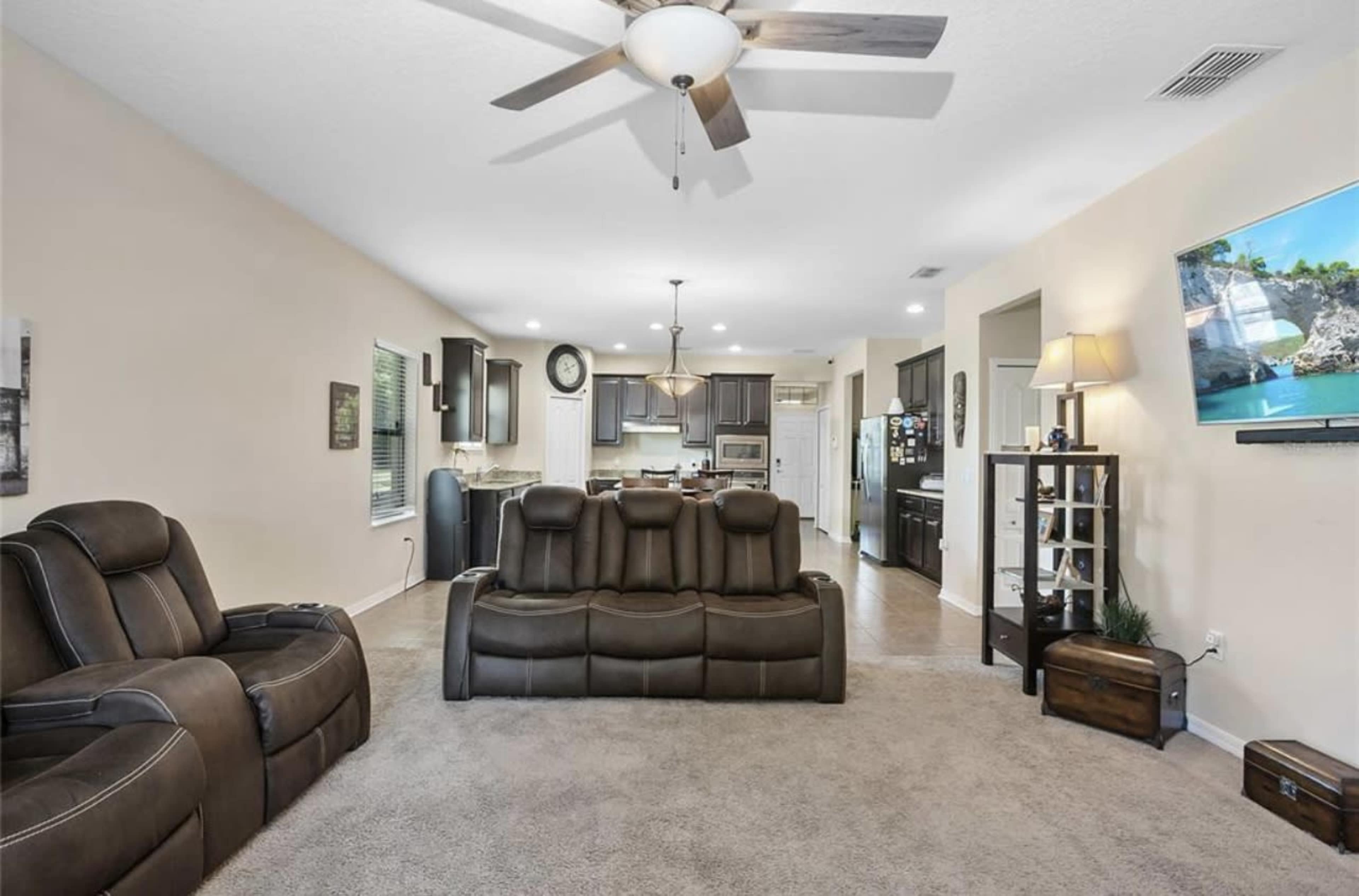 A living room with a dark leather sectional sofa, a ceiling fan, and an open view into a kitchen with dark cabinetry.