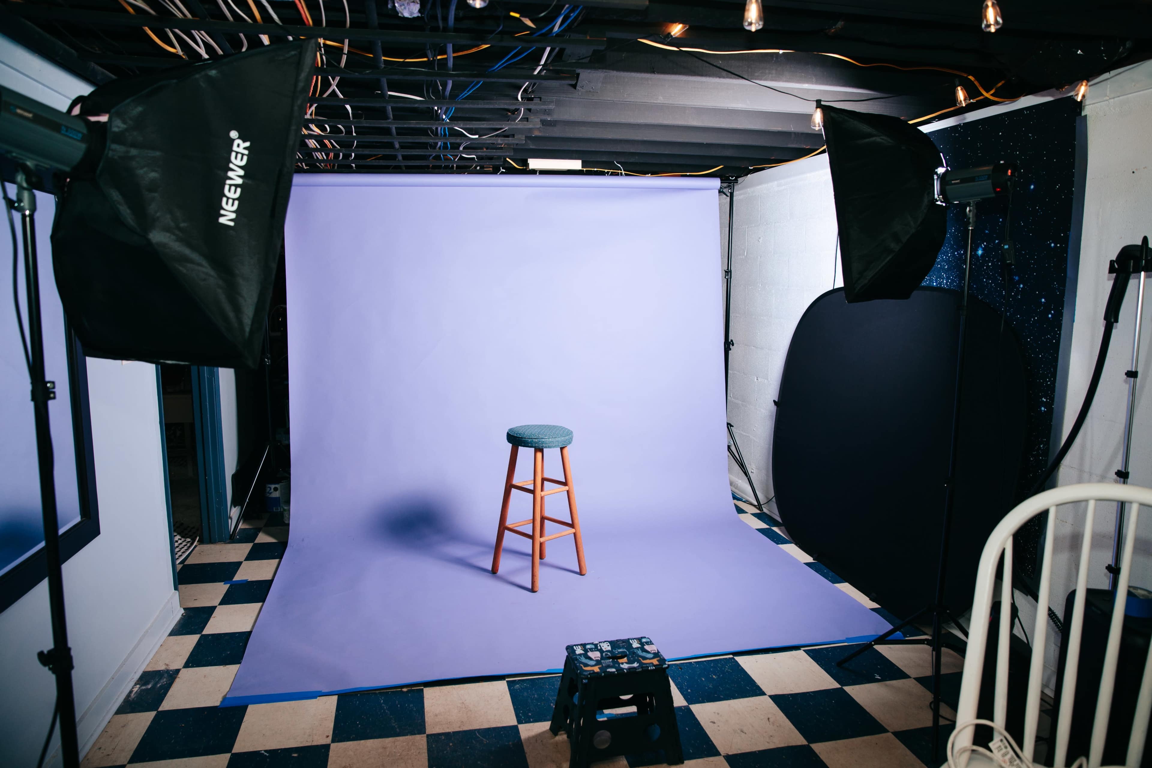 A photography studio is set up with a light purple backdrop, a wooden stool in the center, and studio lighting equipment positioned around the space.