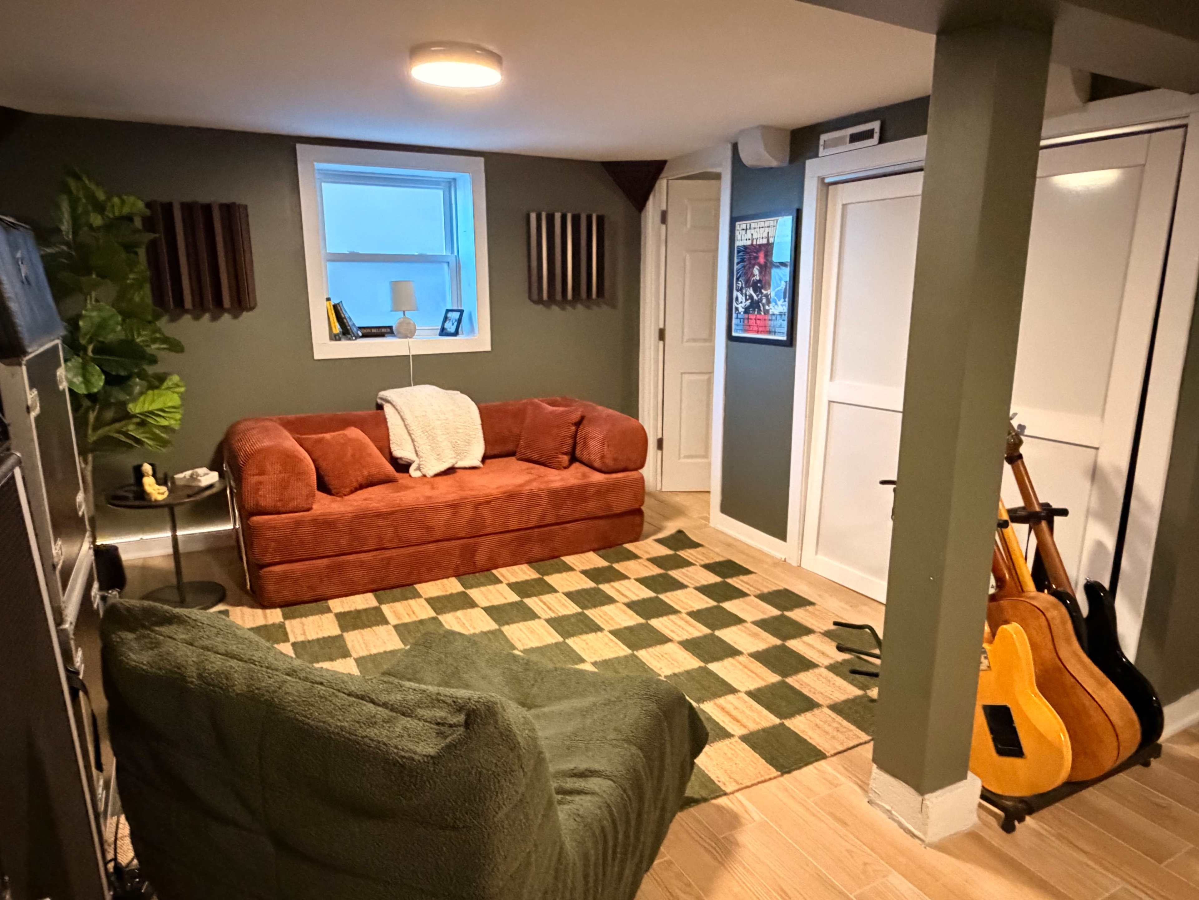 The image shows a cozy basement room with a red couch, green armchair, a checkered rug, and a small bookshelf, along with two acoustic guitars resting against the wall.