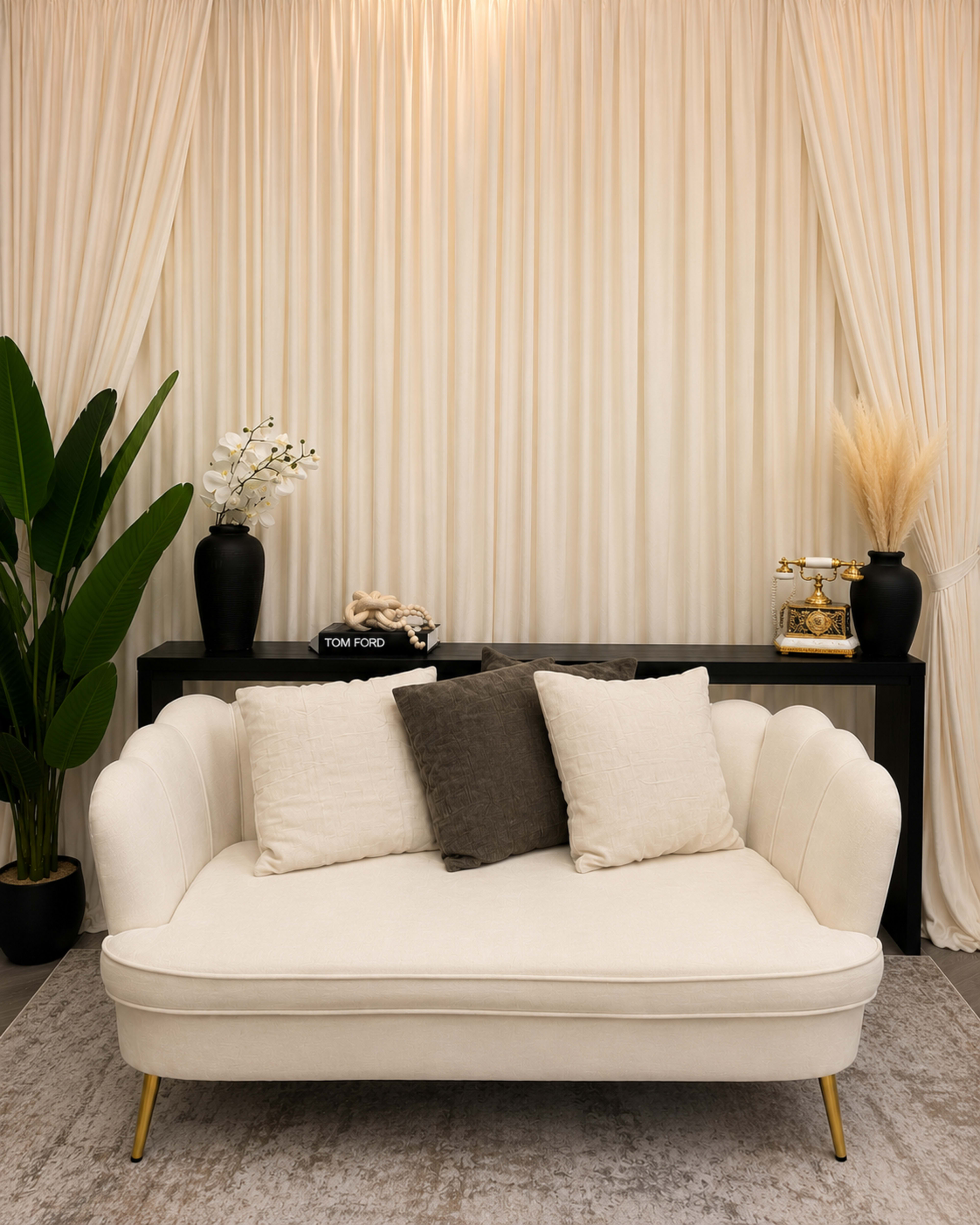 A white, upholstered loveseat with two cushions is placed in front of a black console table and framed by tall, light-colored curtains and green plants.