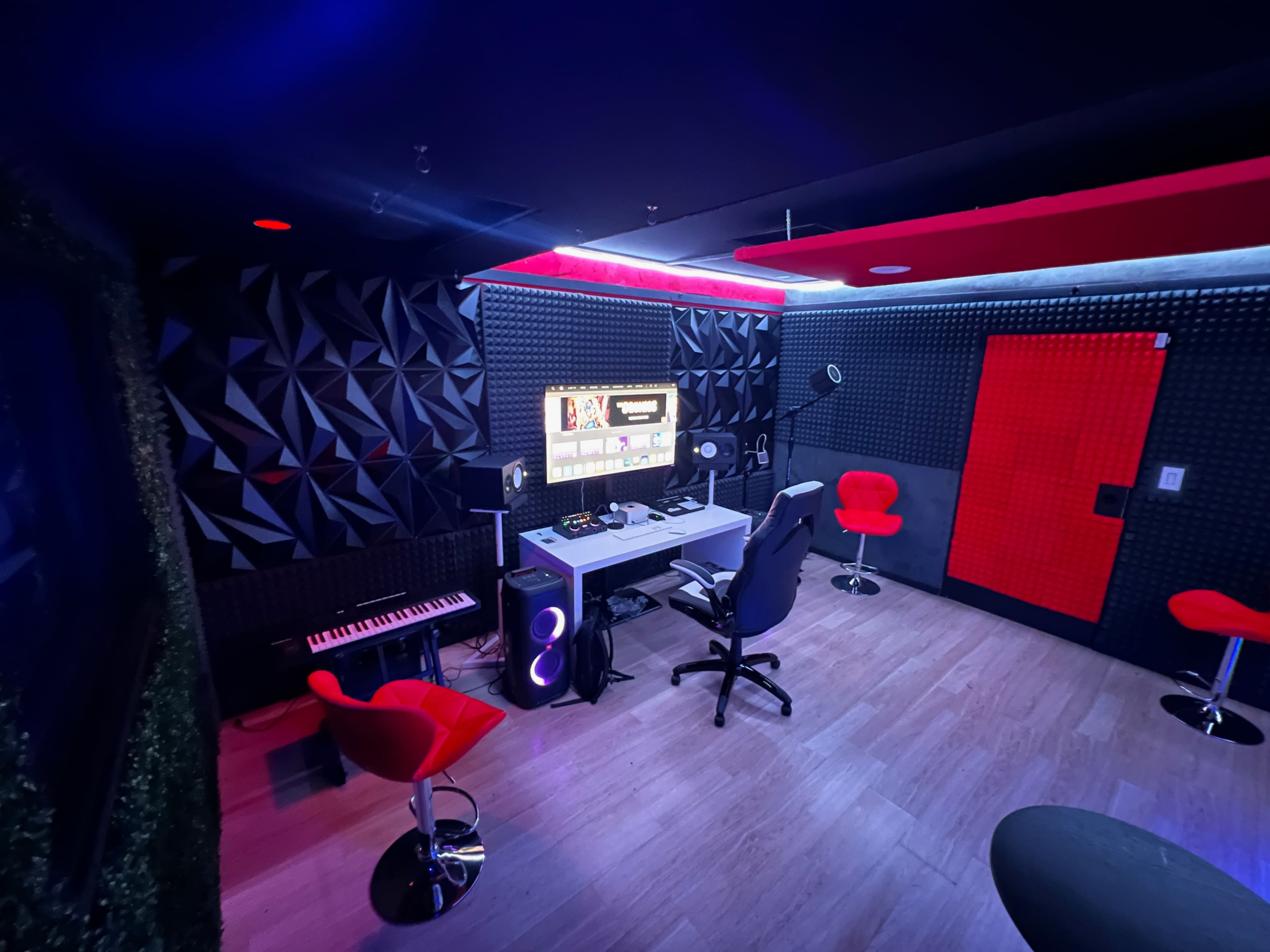 A modern recording studio featuring a central desk with a computer setup, red accent chairs, a keyboard, and soundproof walls.
