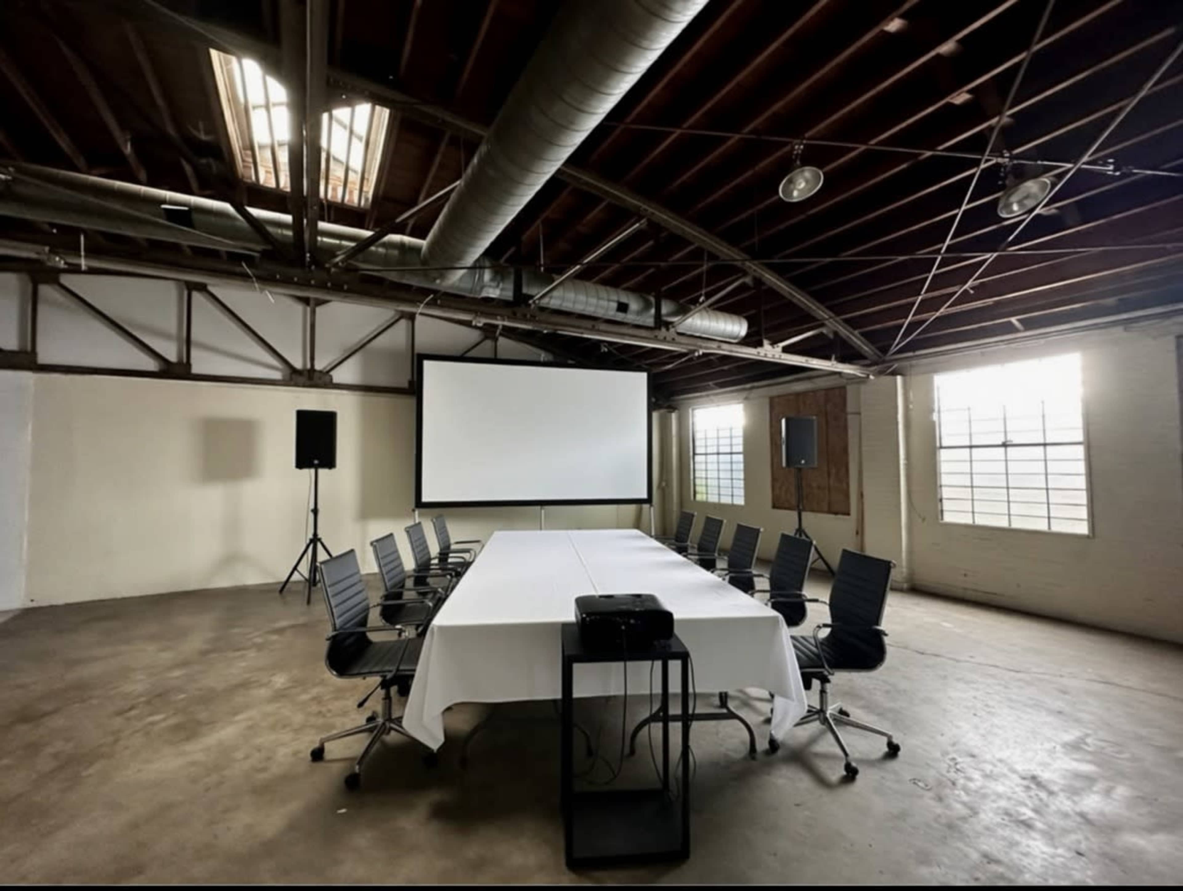 A conference room is set up with a long table, eight chairs, a projector on a stand, and a large screen at one end, all under a high ceiling with exposed beams.