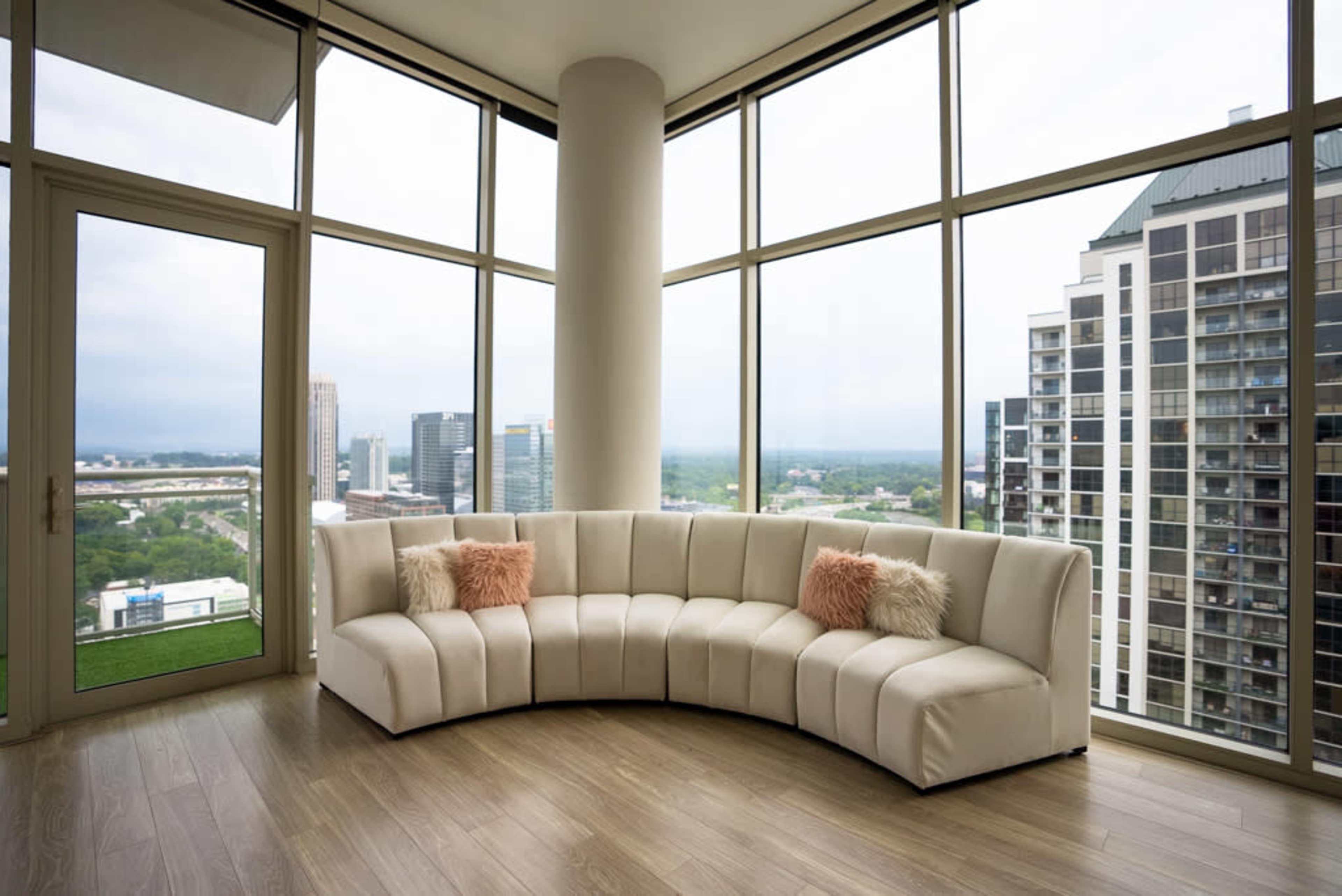 A curved white sofa is positioned in a modern, brightly lit room with large windows offering a view of a city skyline and overcast sky.