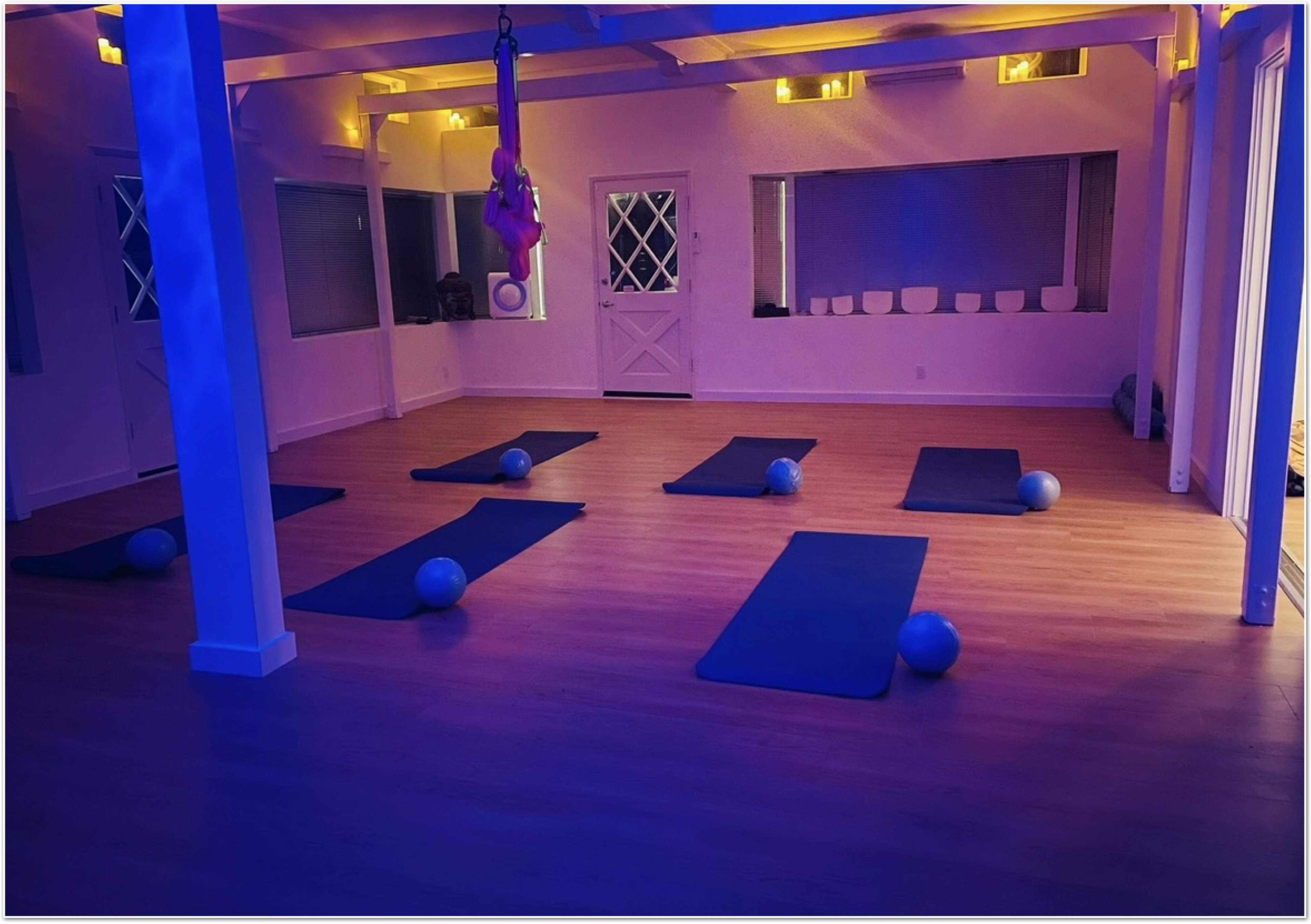 The image shows a fitness studio with several exercise mats and blue exercise balls arranged on a wooden floor, illuminated by soft lighting.
