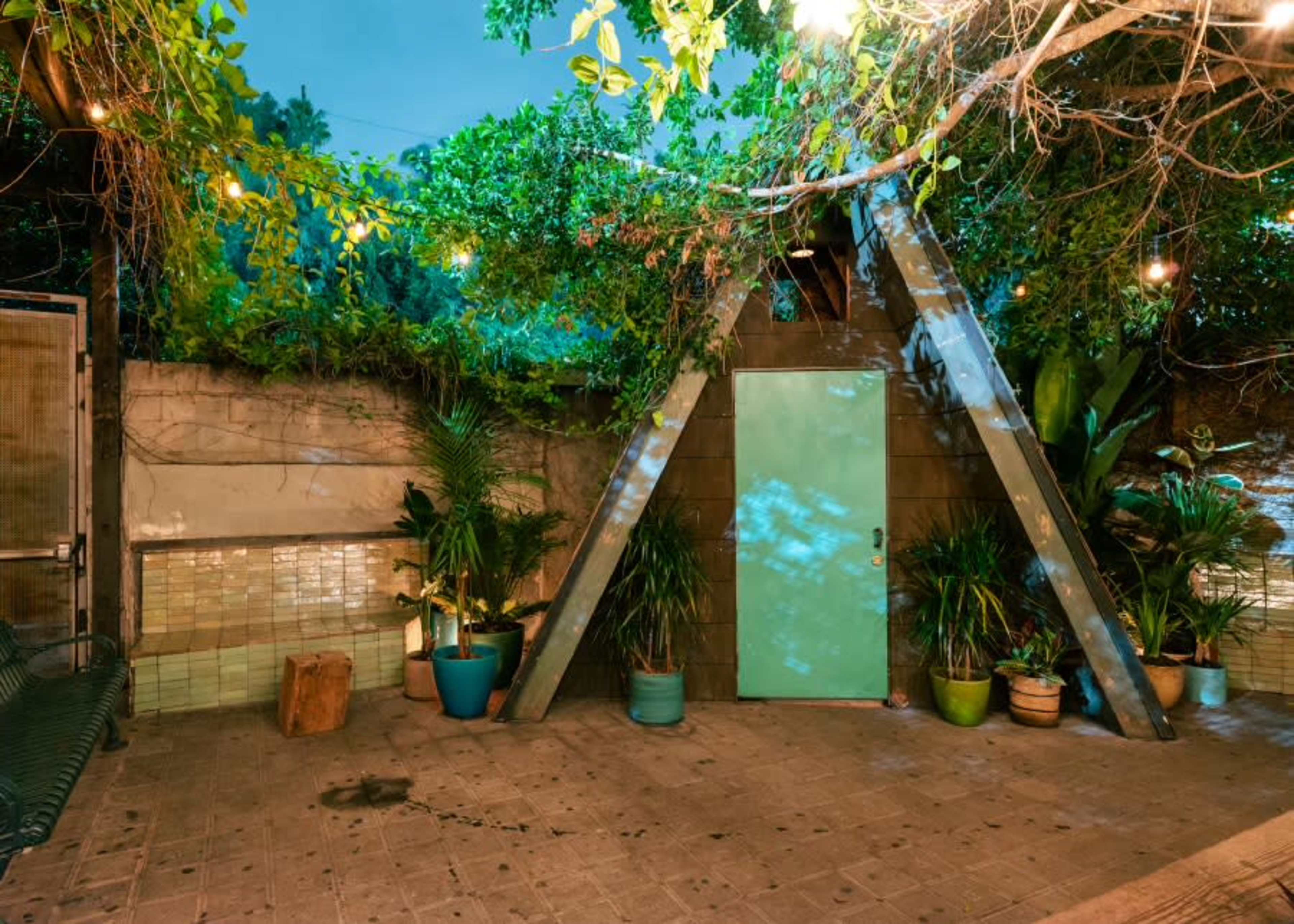 A small, triangular wooden structure stands amidst greenery, with potted plants and string lights illuminating the scene.