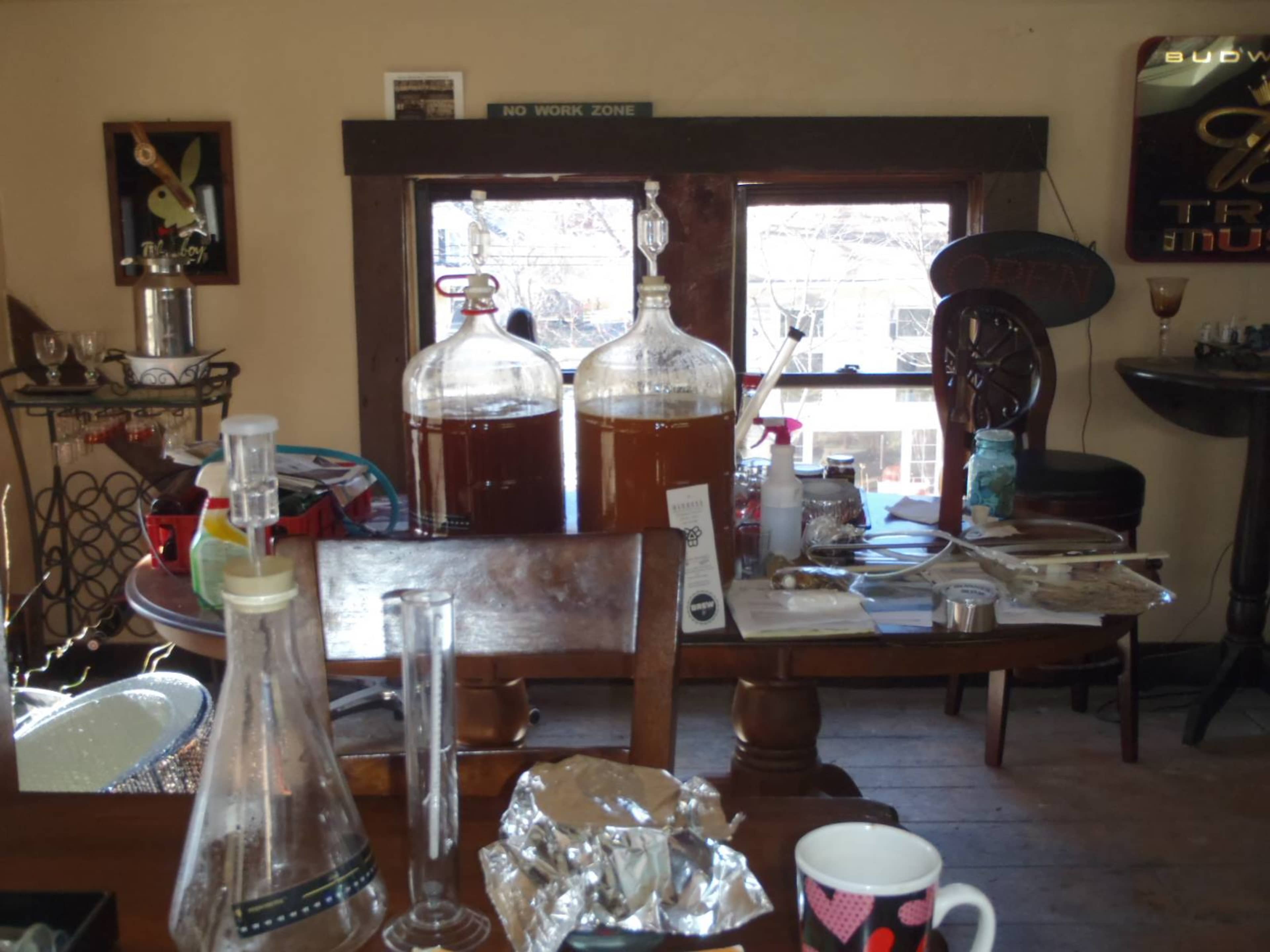 A room with two large glass fermentation vessels, a table cluttered with various items, and a window in the background displaying outdoor scenery.