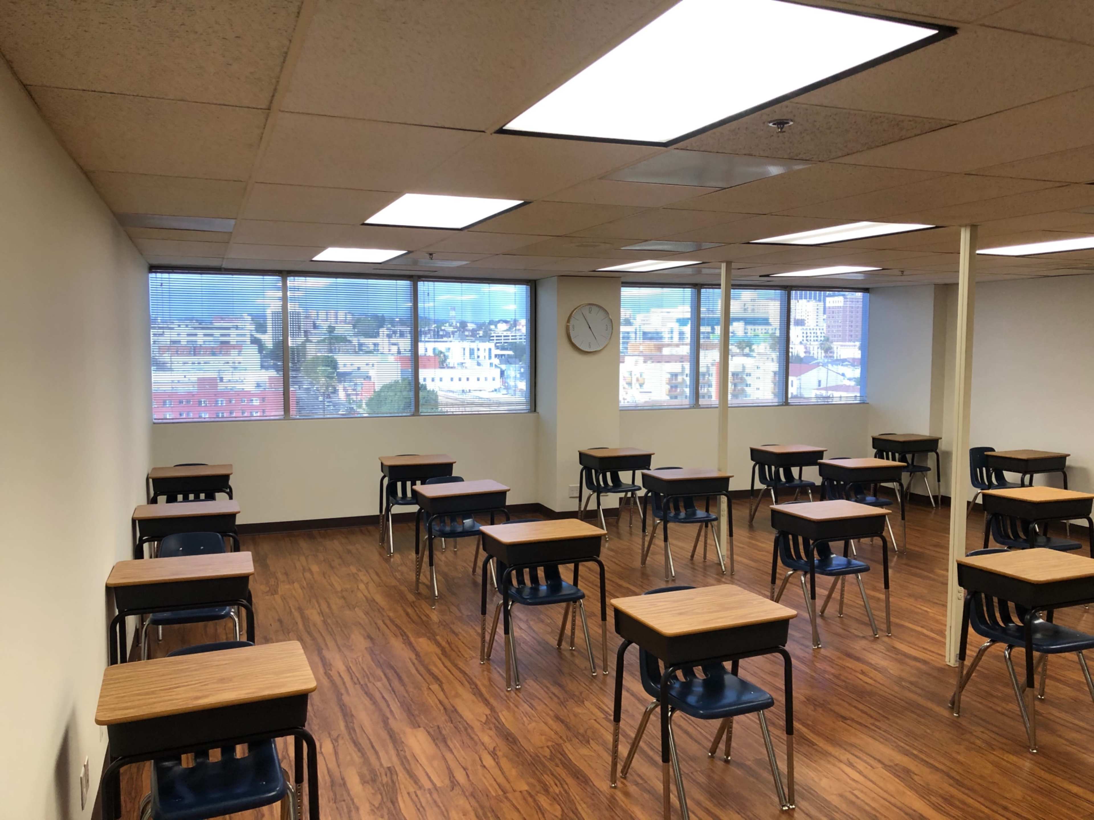 The image shows a classroom with empty desks arranged in a grid pattern and large windows providing a view of an urban landscape.