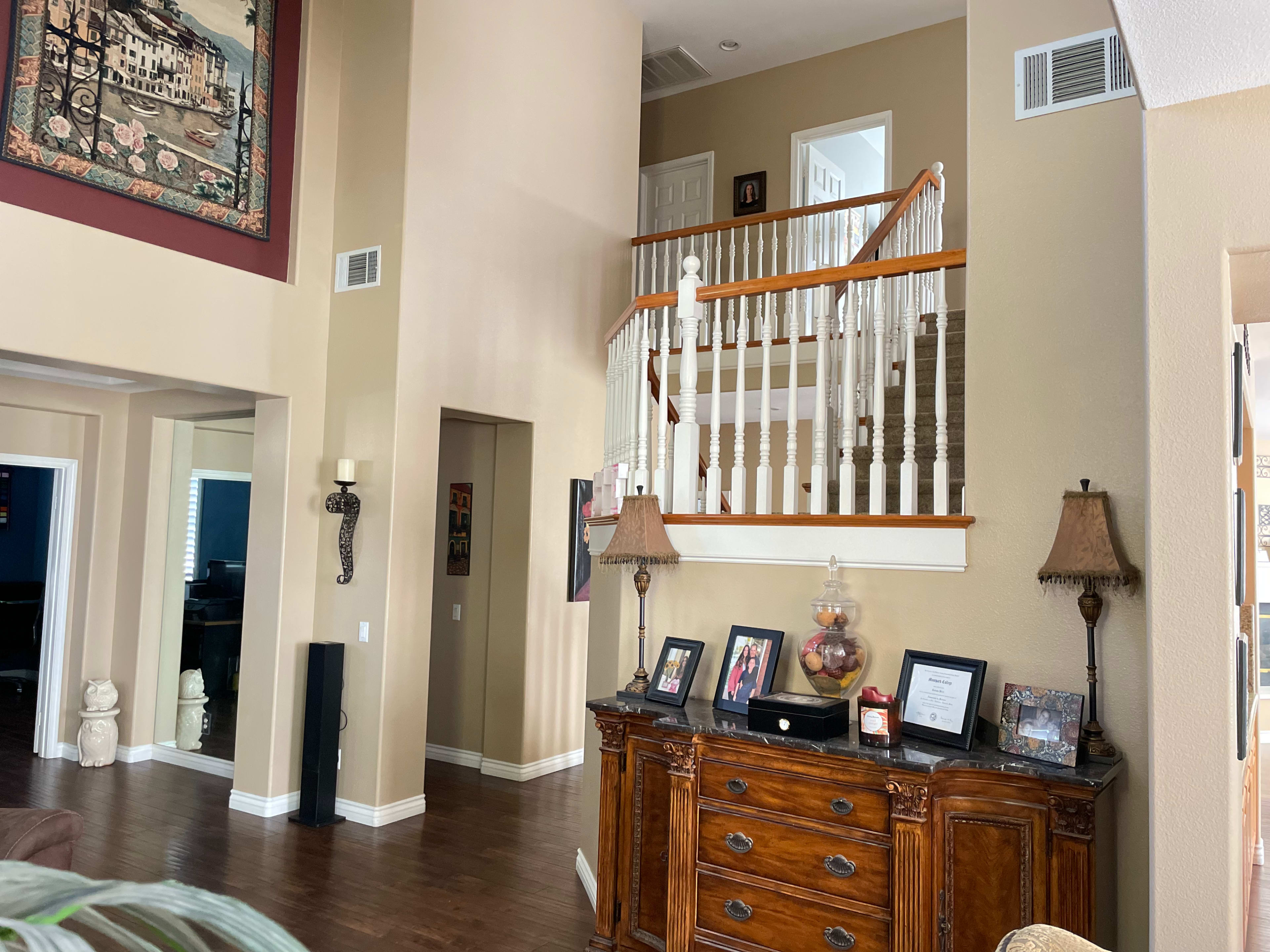 A spacious living area with wooden flooring, featuring a staircase in the background and a decorative cabinet with family photos and lamps in the foreground.