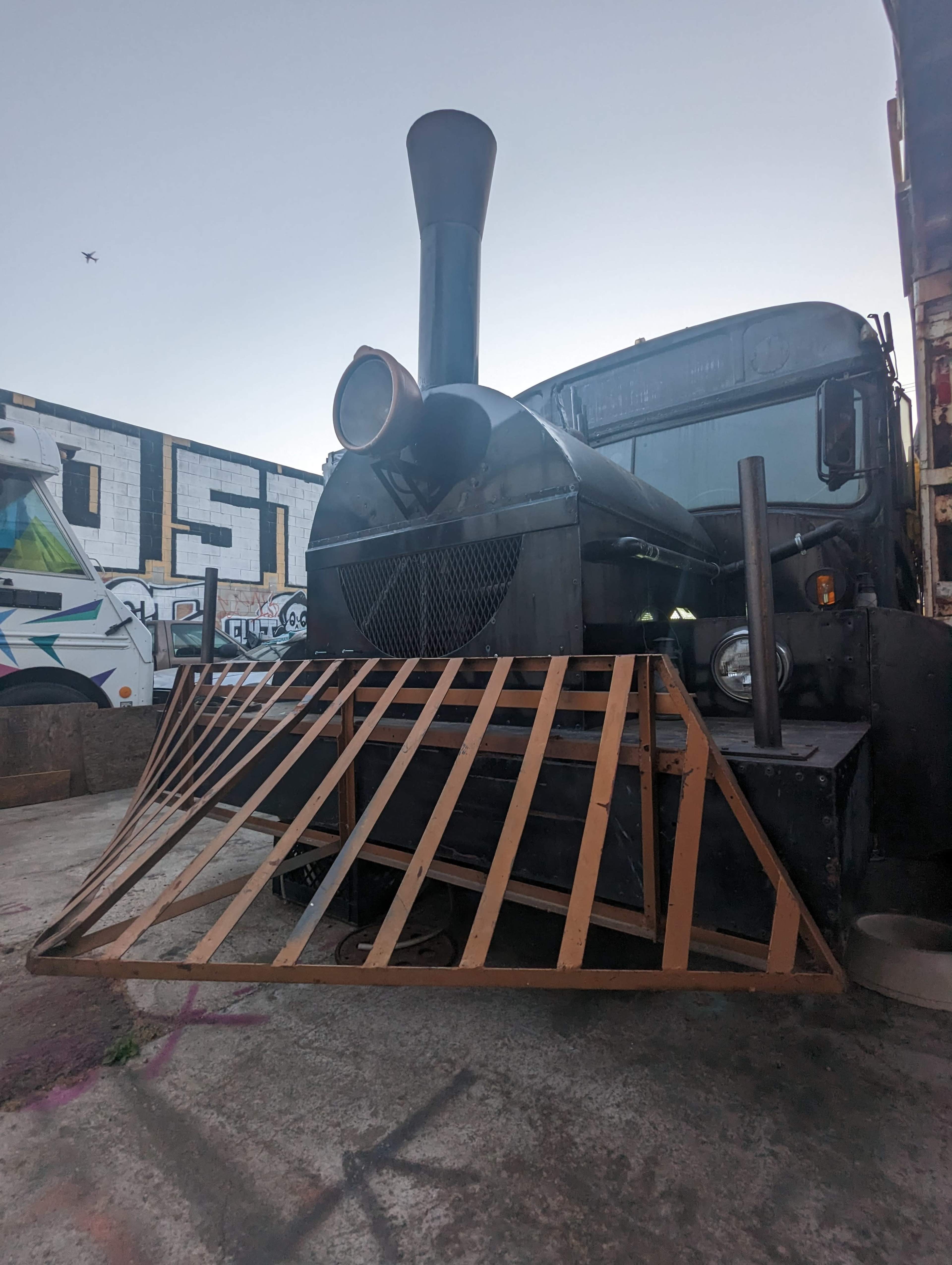 A vintage truck with a large steam engine attachment and a wooden ramp is parked in an industrial area.
