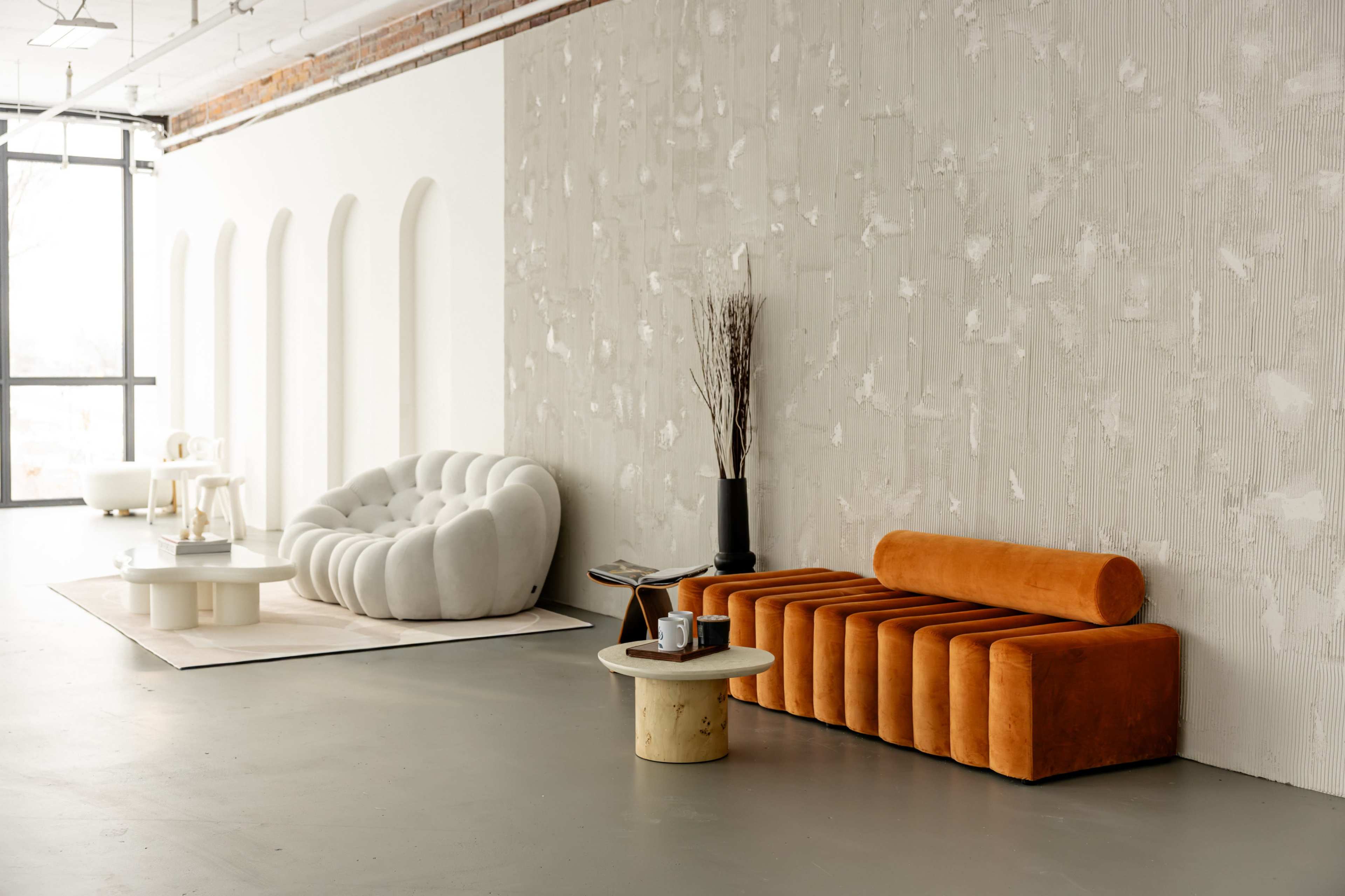 The image shows a modern interior with a textured gray wall, a curved white sofa, an orange velvet bench, and a round coffee table positioned on a light-colored rug.