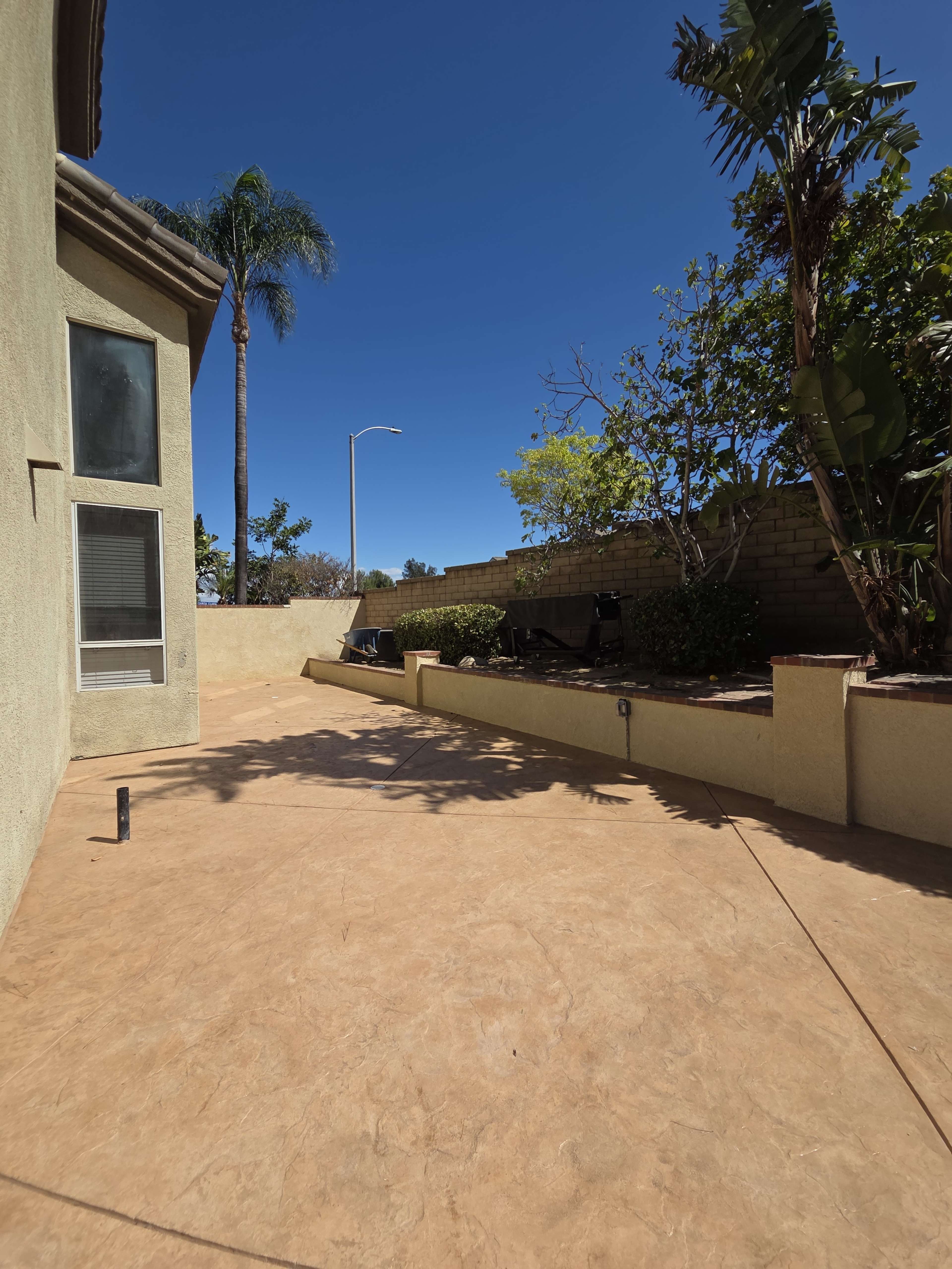 The image shows a large, empty patio area next to a house, with a clear blue sky and a few plants and trees in the background.