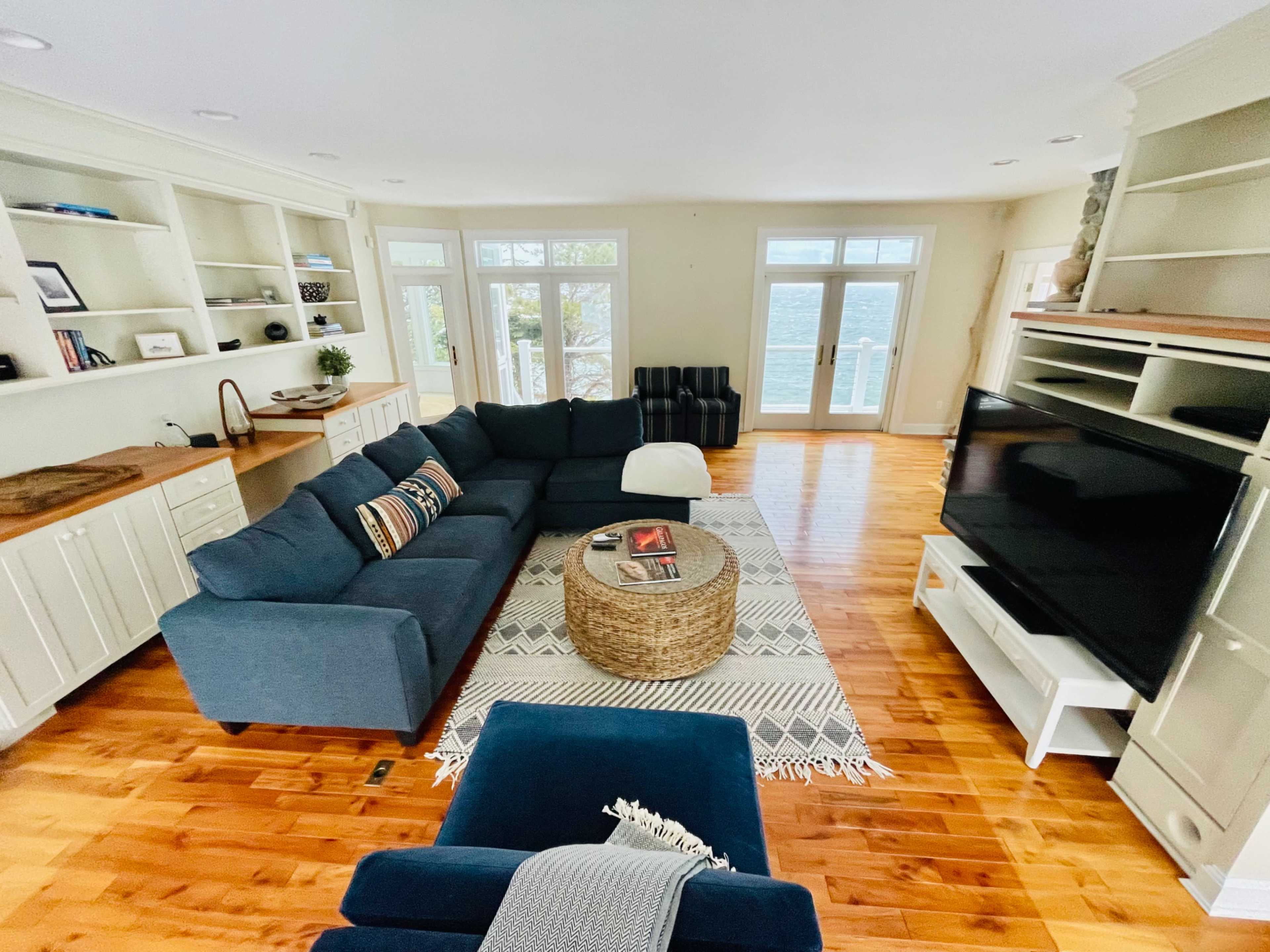 A spacious living room with a large blue sectional sofa, a round coffee table, and a television against a cream-colored wall, illuminated by natural light from large windows.
