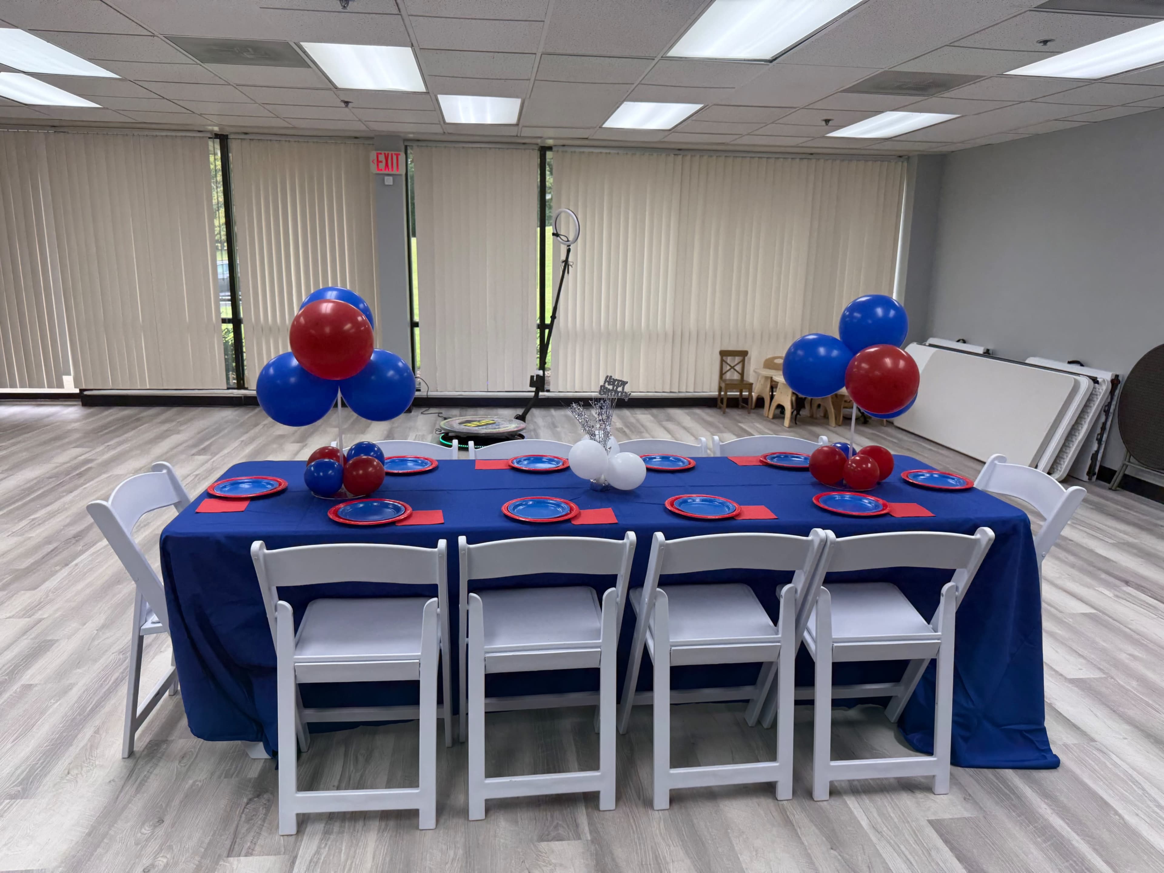 A decorated table with blue and red tablecloths is set up with plates and balloons, arranged in a room with large windows and a wooden floor.