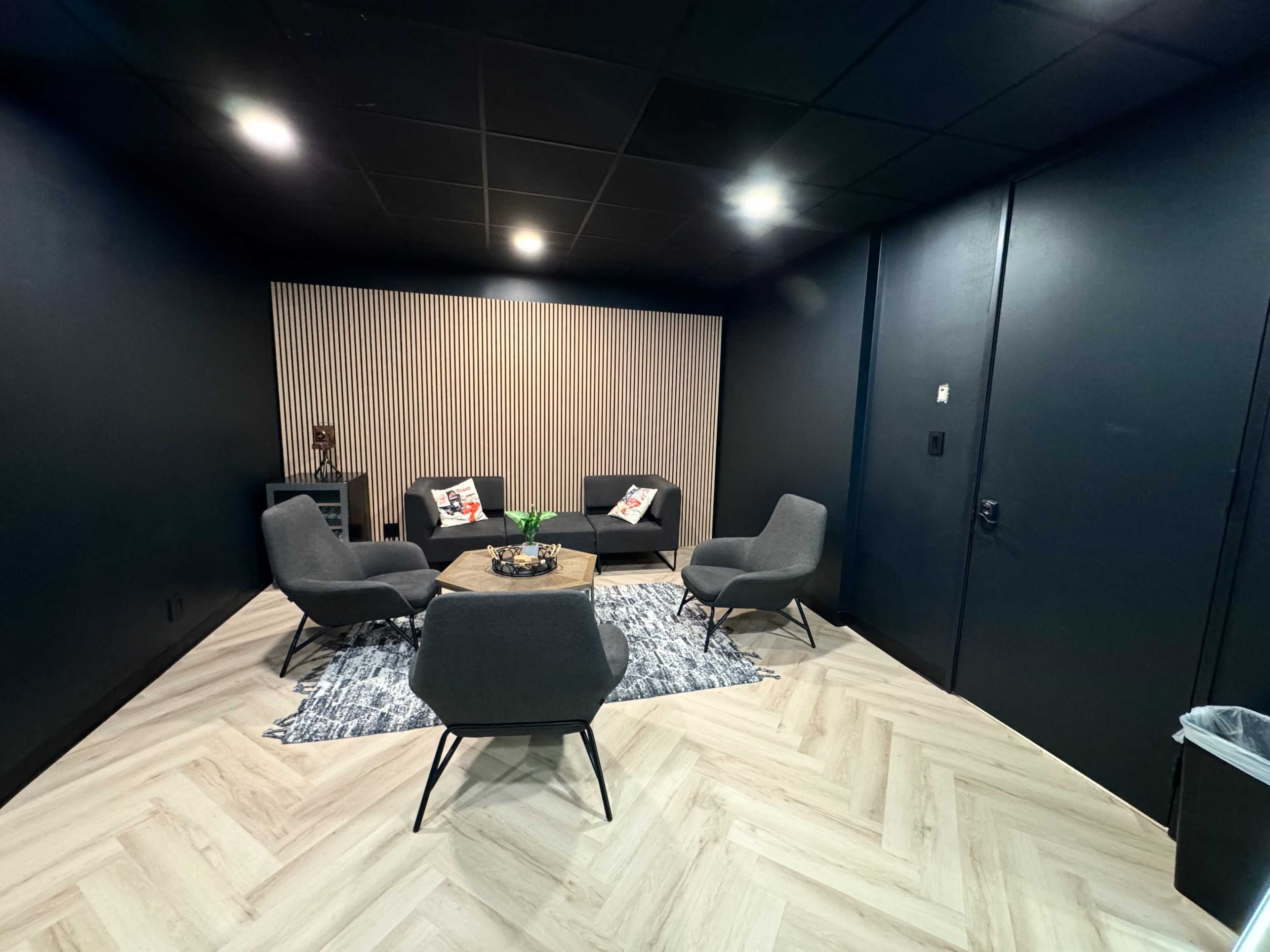 The image shows a small meeting room featuring four gray chairs arranged around a circular wooden table on a patterned rug, with a wooden accent wall in the background.