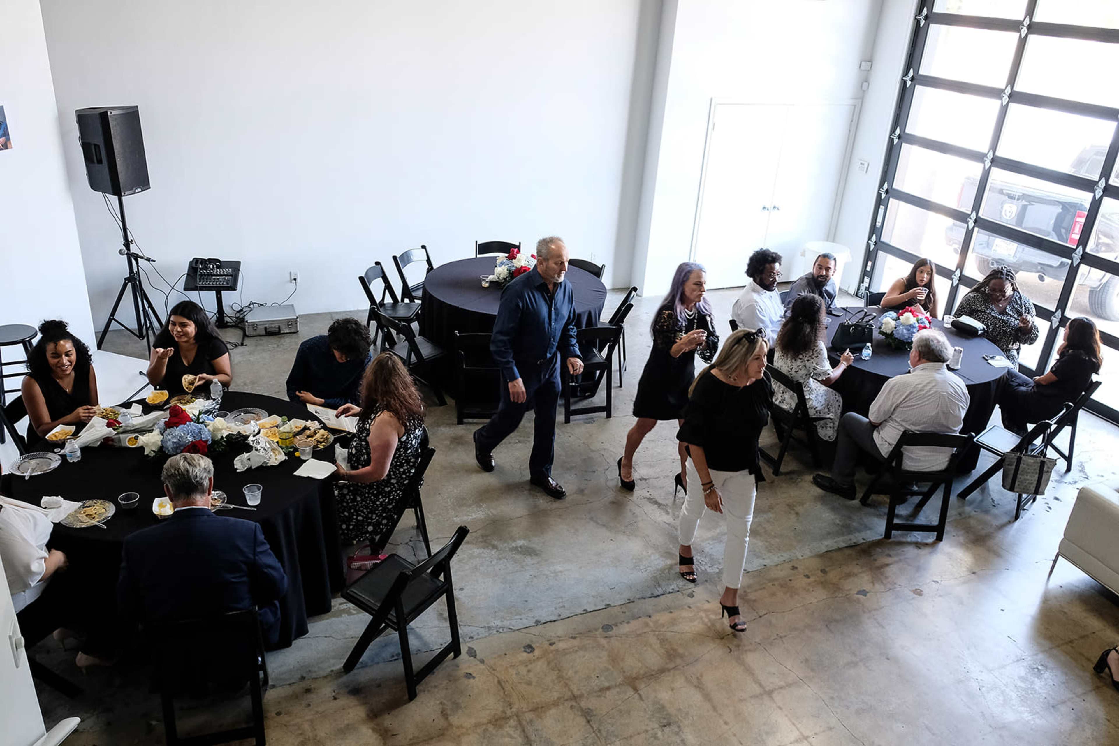 A group of people are gathered at tables in a bright event space, enjoying food and drinks while some mingle.