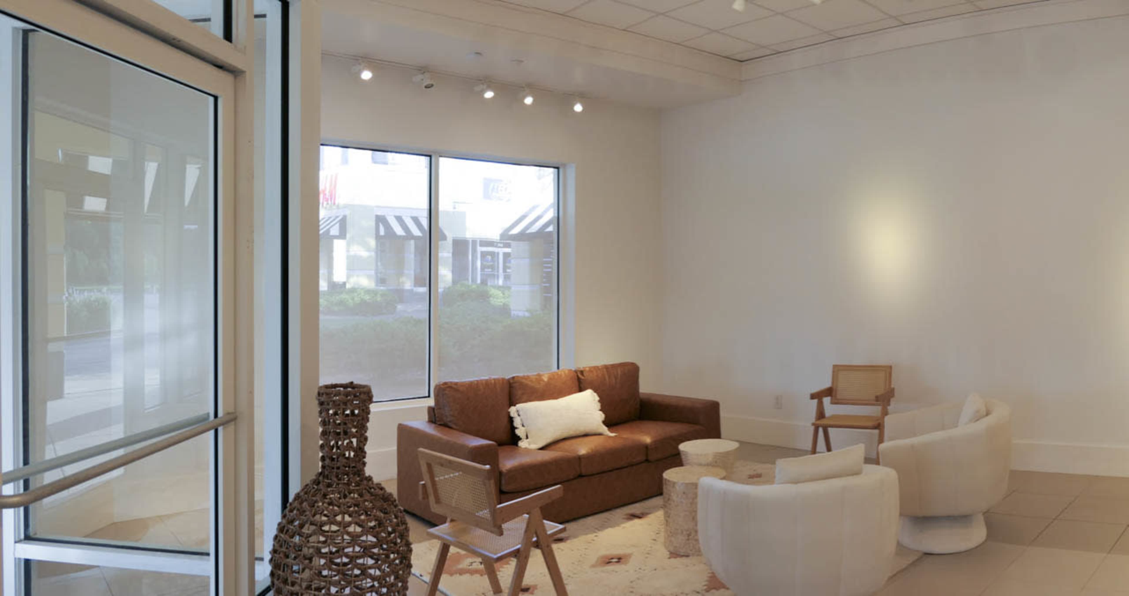 The image shows a modern, minimalist waiting area with a brown leather sofa, two white armchairs, a light-colored rug, and a decorative woven sculpture.