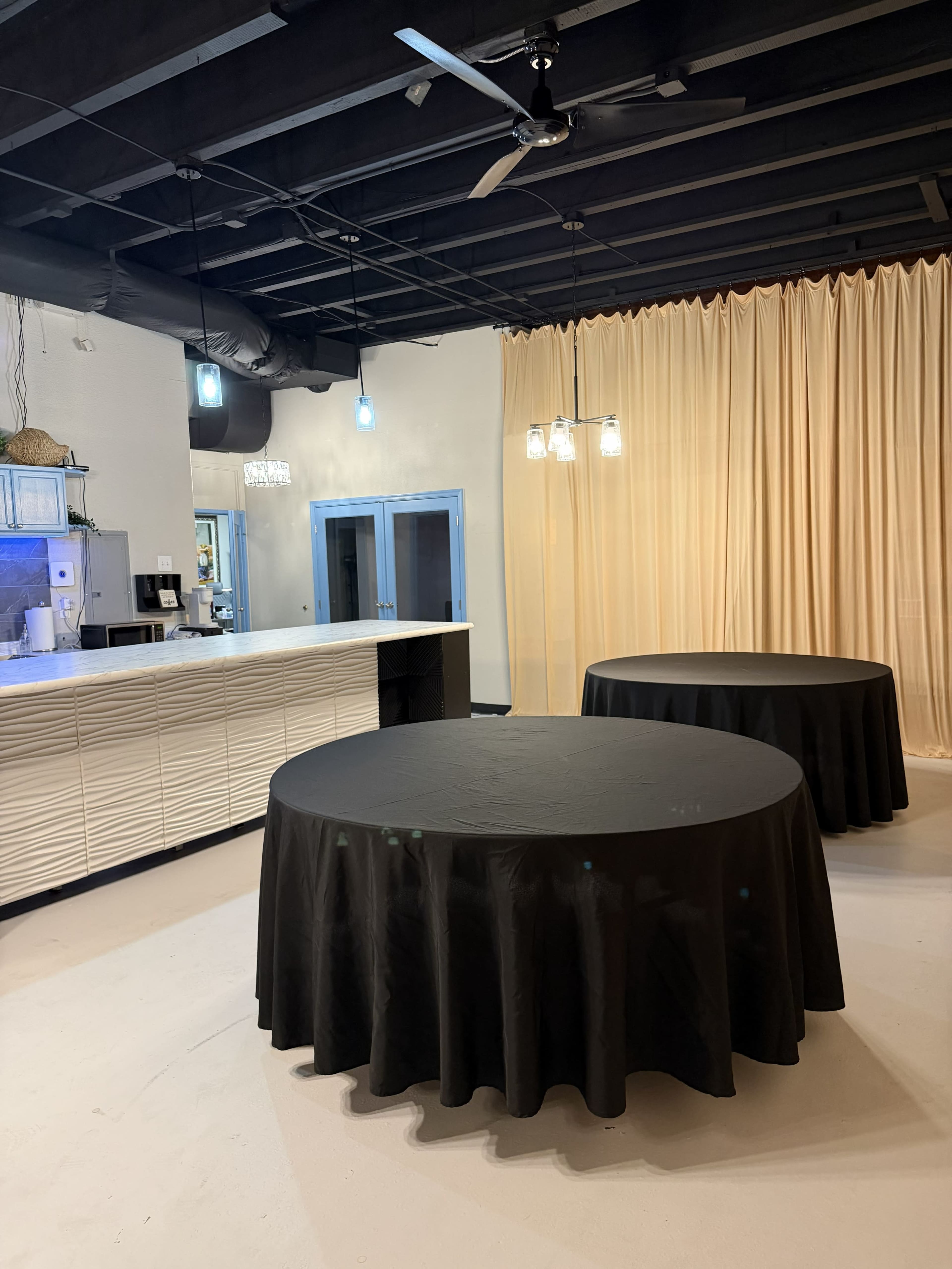 The image shows an interior space featuring two round tables covered with black tablecloths, a light-colored counter, and a draped curtain along one wall.