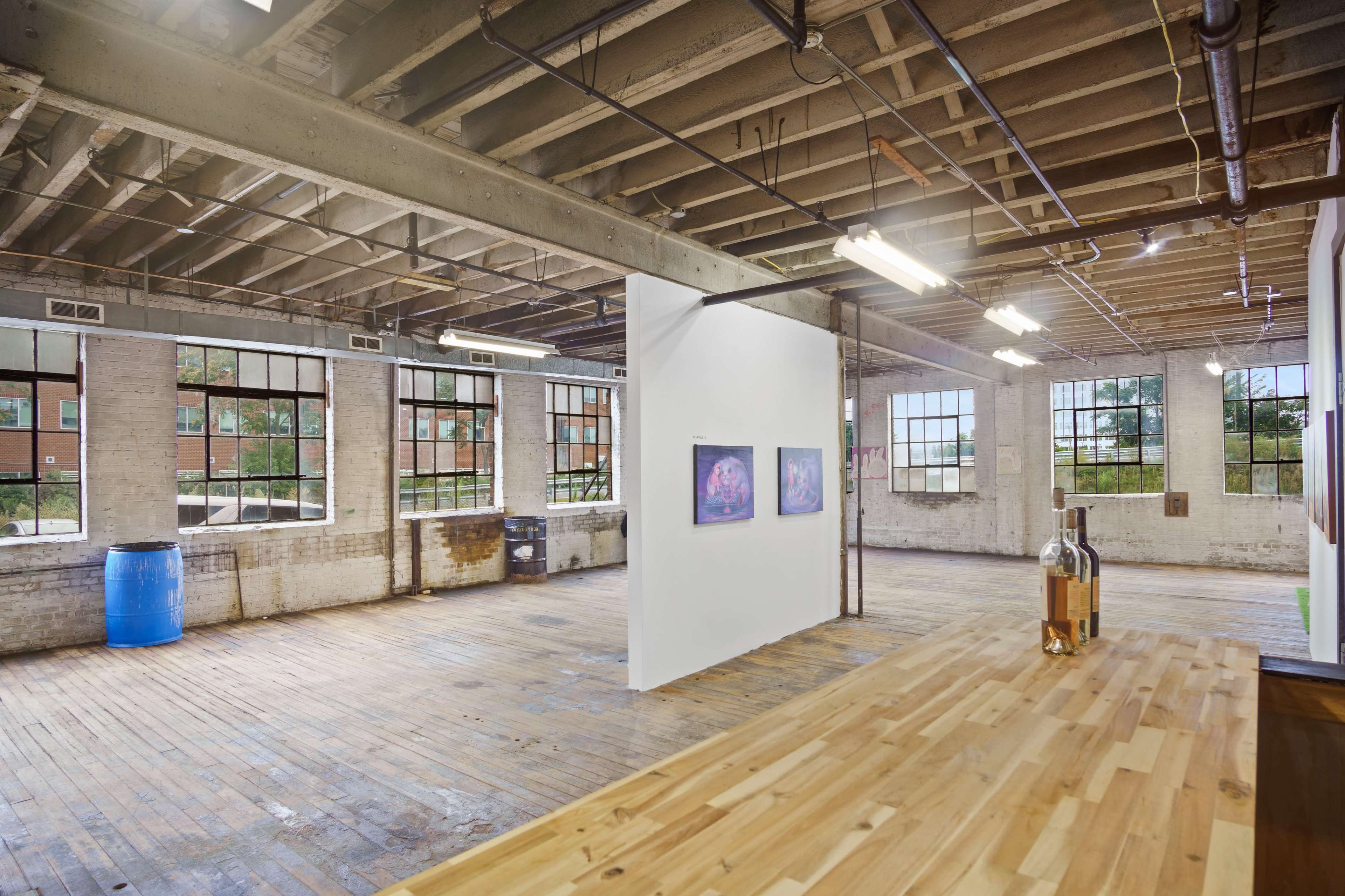 A spacious, industrial-style art gallery featuring wooden floors, large windows, and a dividing wall with framed artwork.
