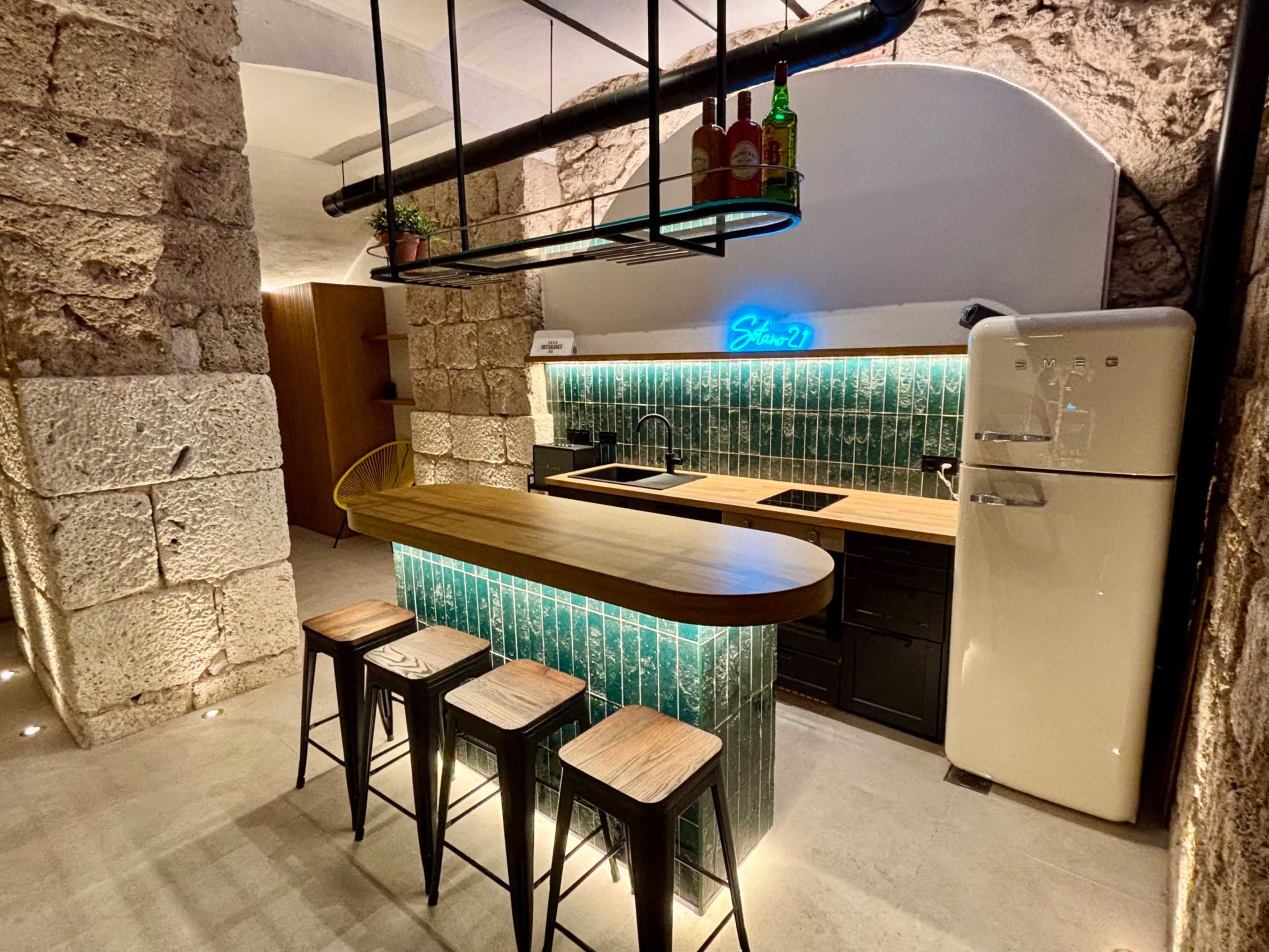 The image shows a modern kitchen featuring a wooden island with bar stools, a vintage-style refrigerator, and a backsplash of green tiles illuminated by blue lighting.