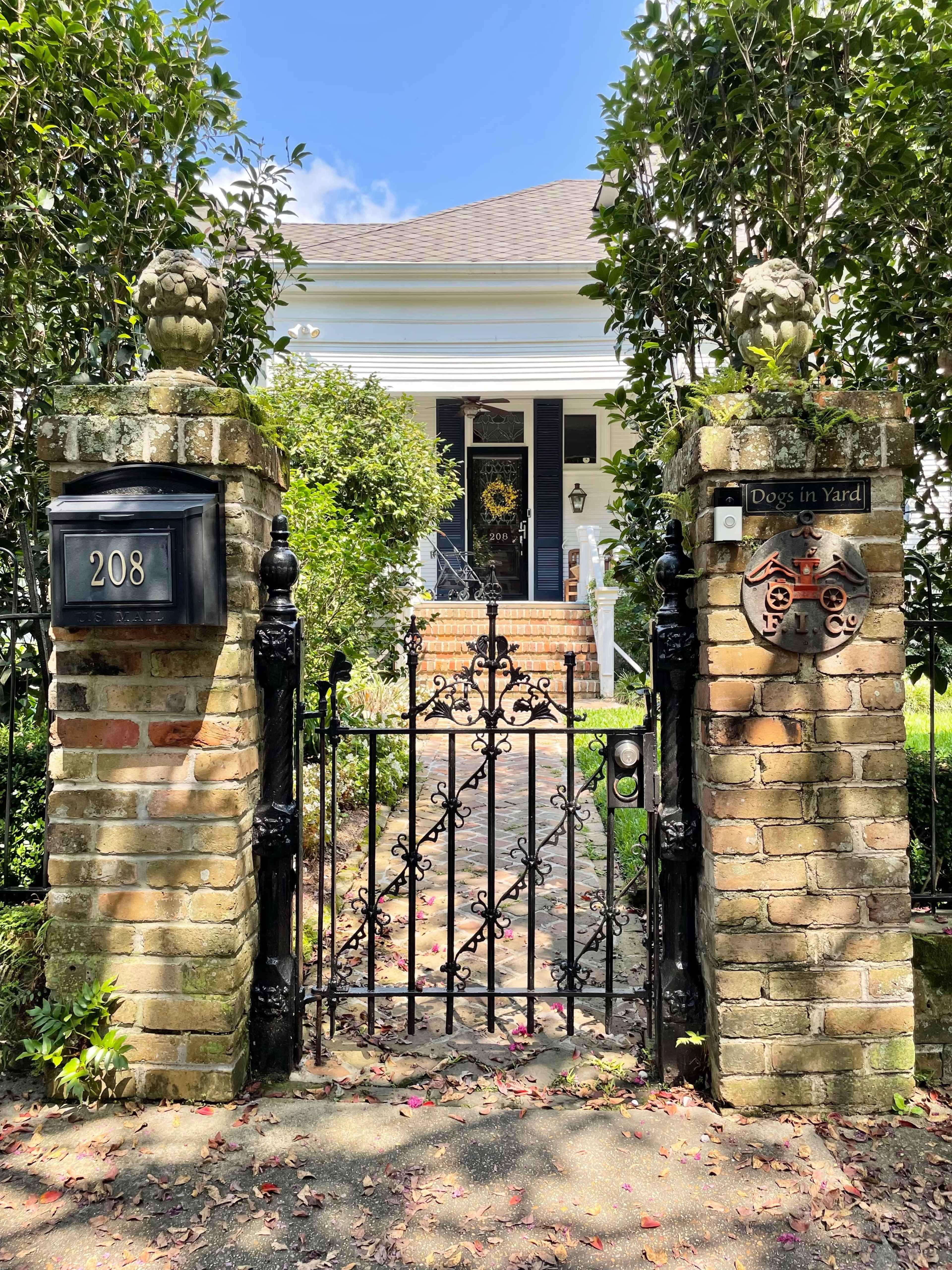 A wrought iron gate with decorative stone pillars leads to the front steps of a white house surrounded by green foliage.