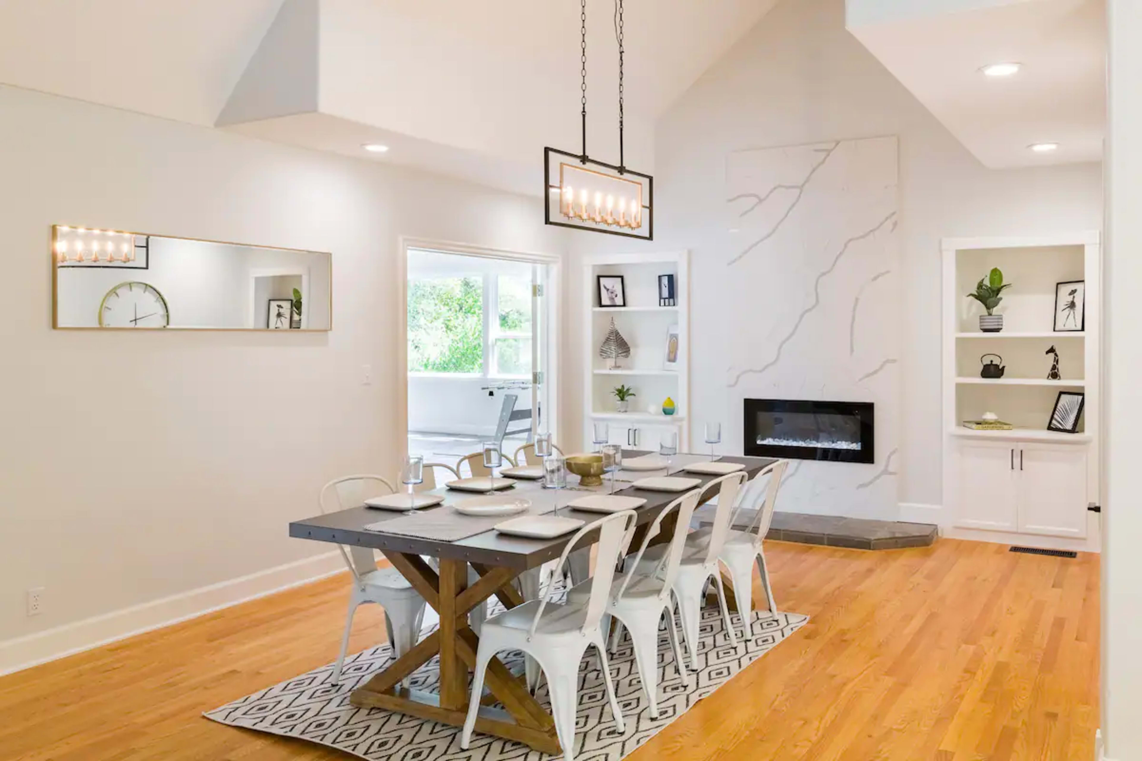 A spacious dining area features a large wooden table with eight chairs, a modern light fixture above, and a wall-mounted fireplace surrounded by decorative shelving.