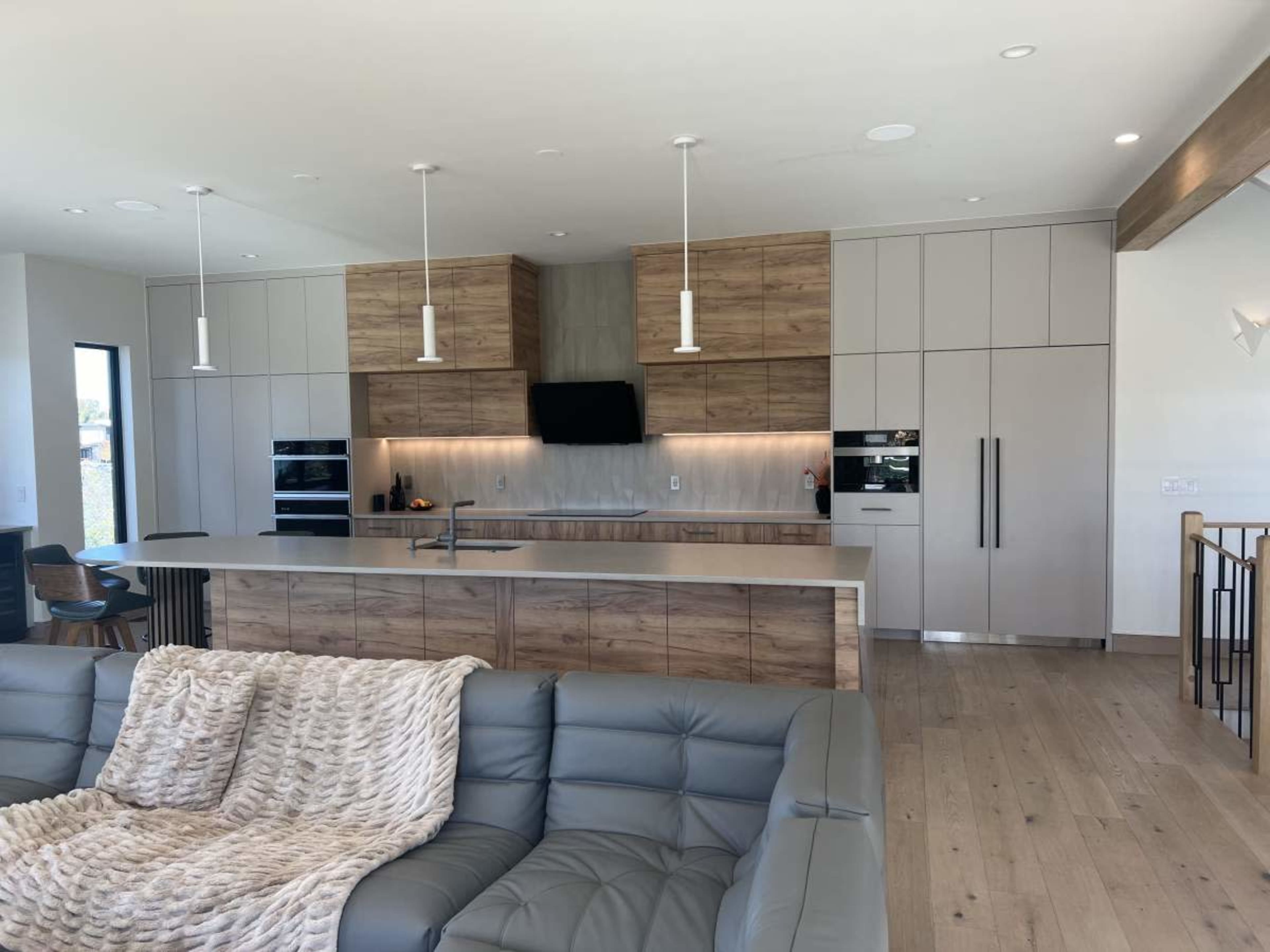 The image shows a modern kitchen with wood cabinetry, a gray countertop, and stainless steel appliances, adjacent to a sitting area furnished with a gray couch.