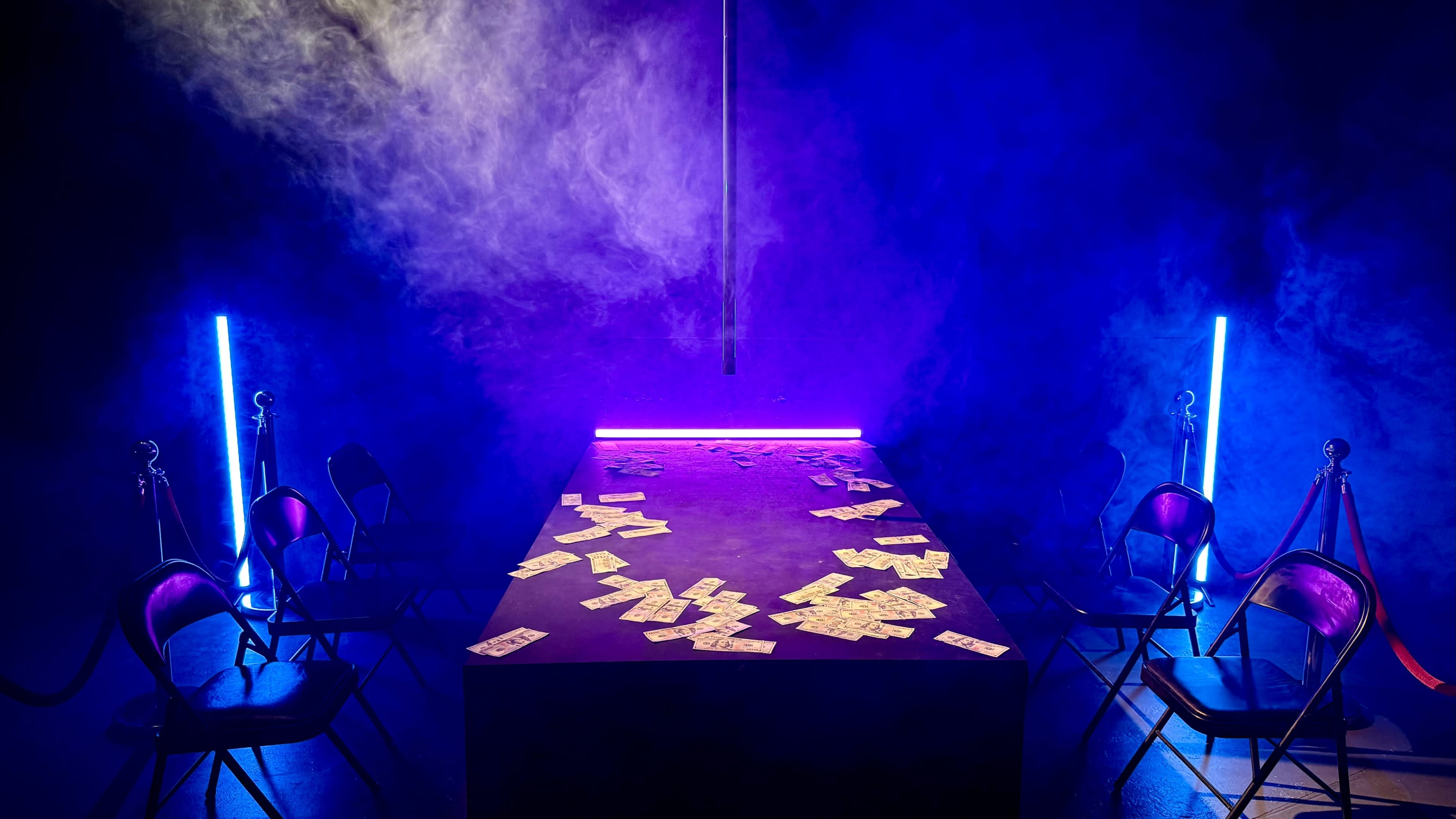 A dimly lit room features a table scattered with dollar bills, surrounded by empty chairs and illuminated by neon lights.