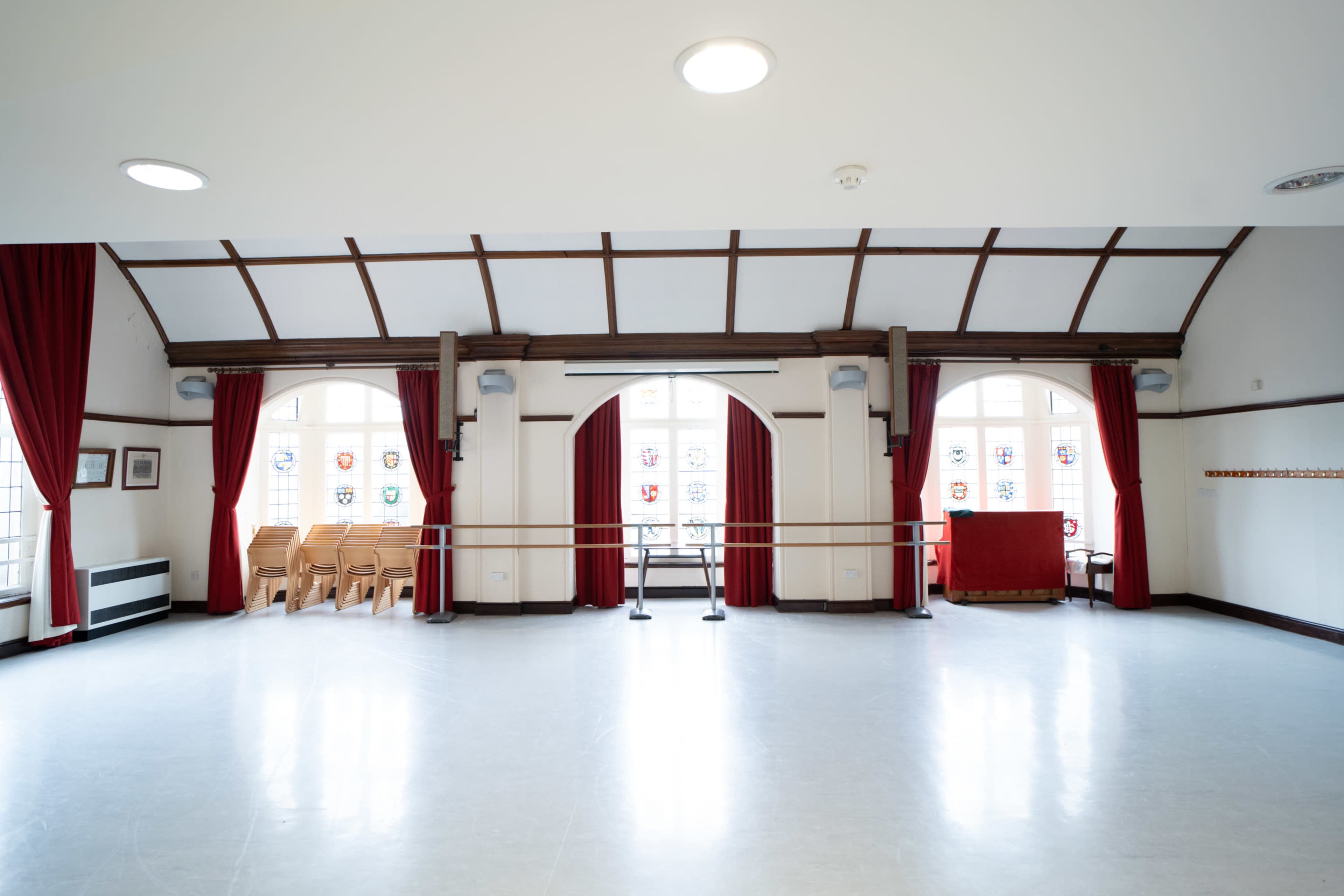 The image shows a spacious dance studio with wooden accents, large windows, and red curtains, featuring a ballet barre and rows of chairs stacked against the wall.
