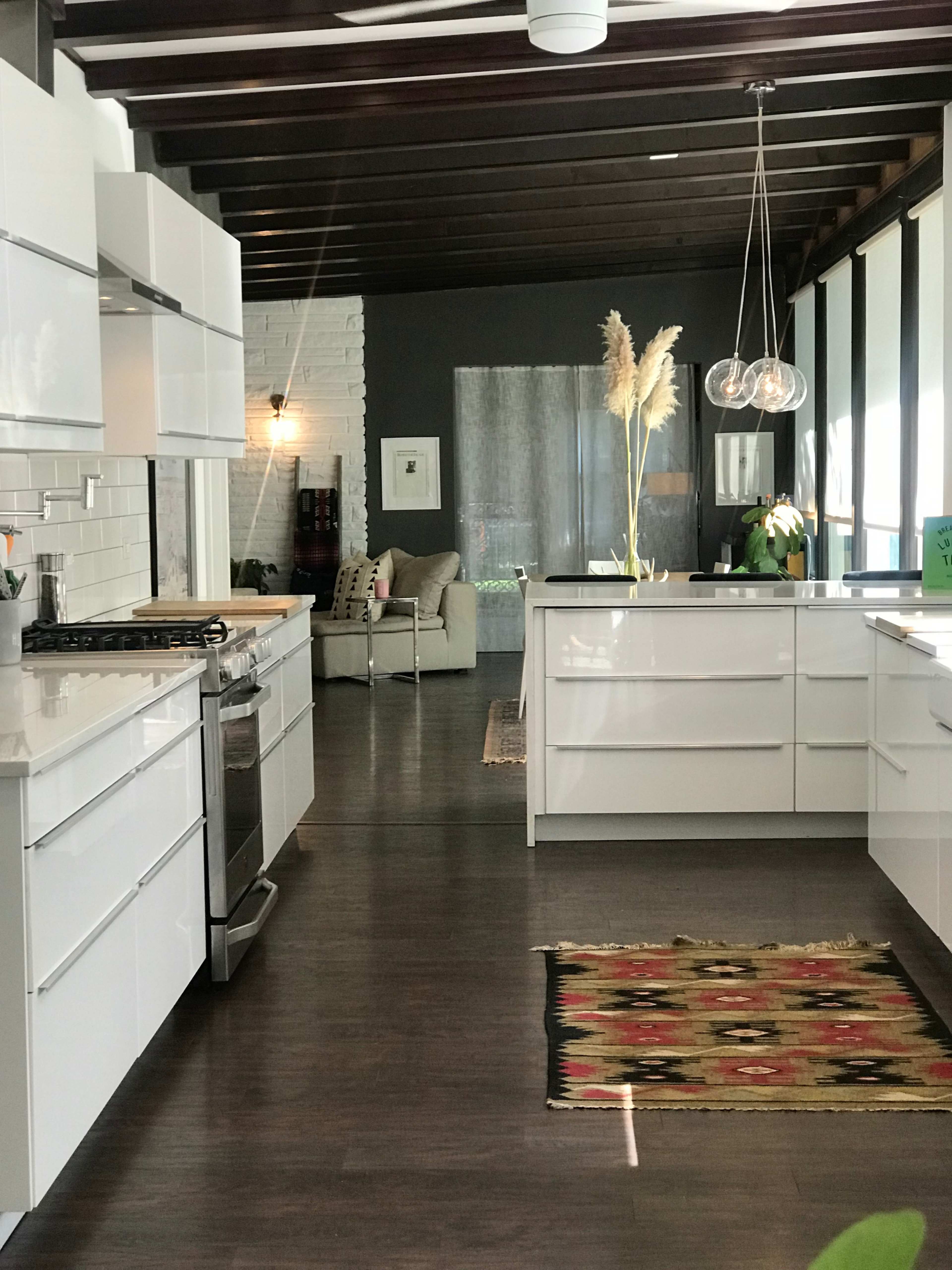 A modern kitchen features white cabinetry, a stainless steel stove, and hardwood floors, with a living area visible through large windows.