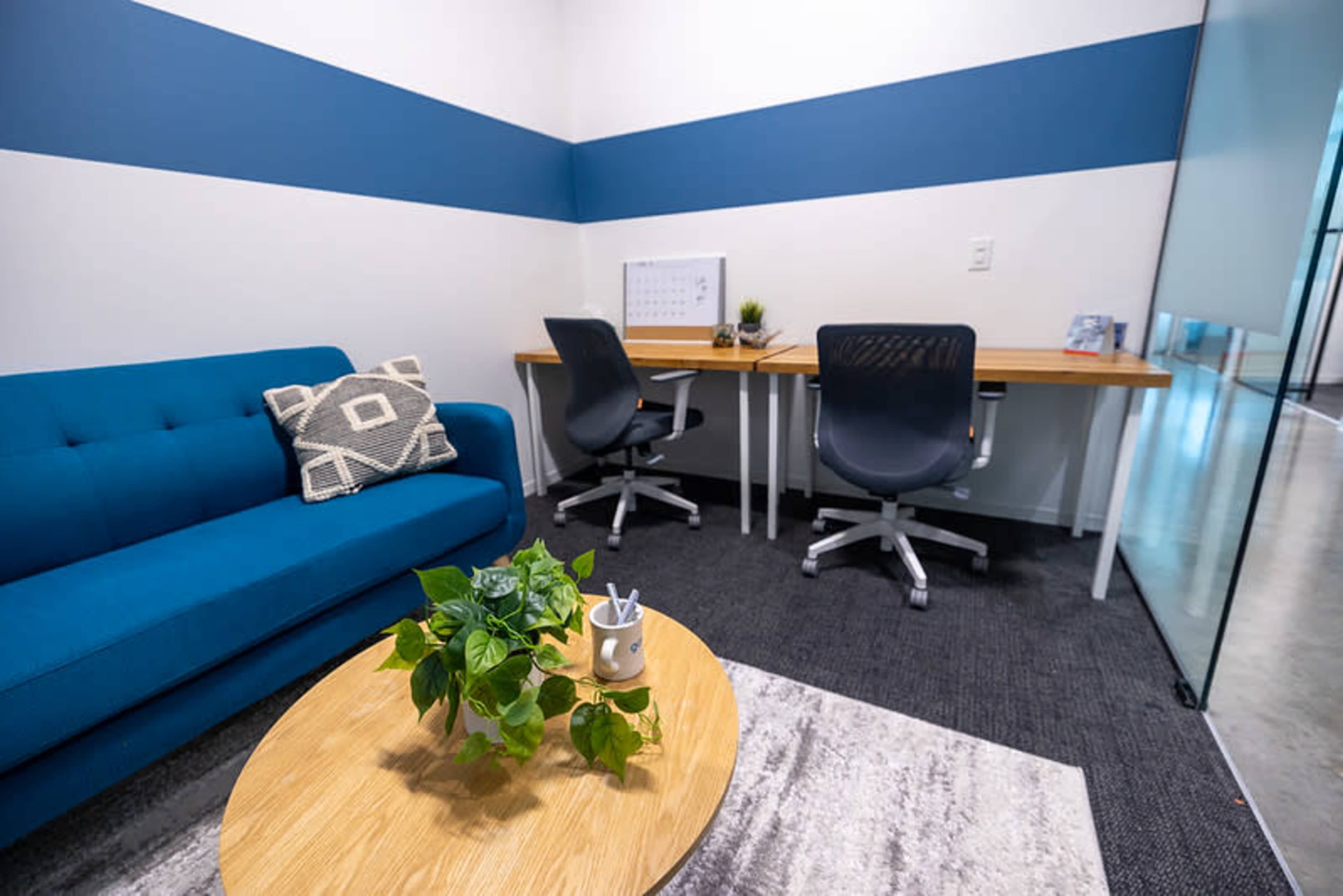 The image shows a small office space with a blue couch, a circular wooden coffee table with a plant, and two ergonomic chairs placed at a desk.