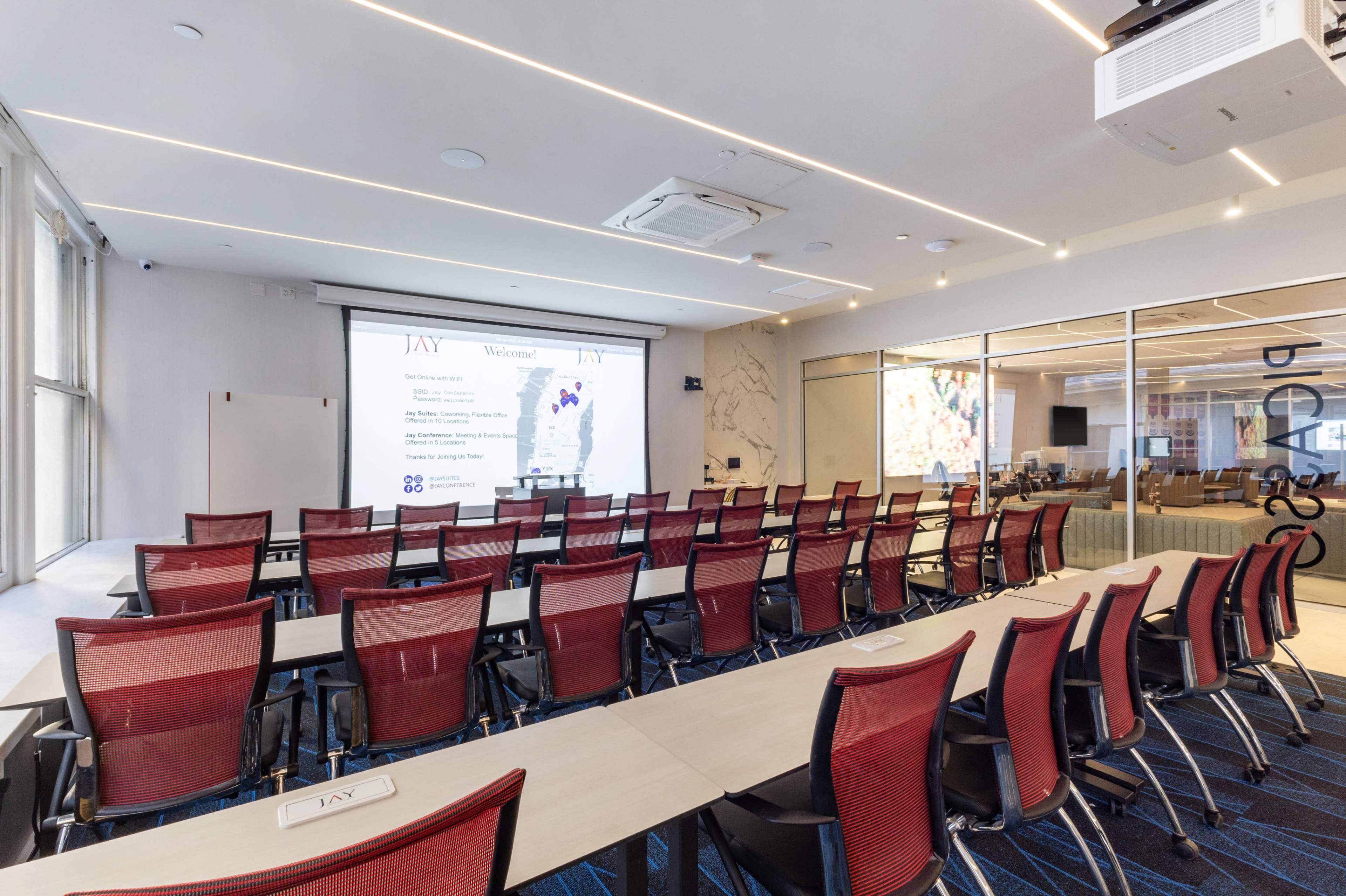 The image shows a modern conference room with rows of red mesh chairs facing a screen displaying a presentation.