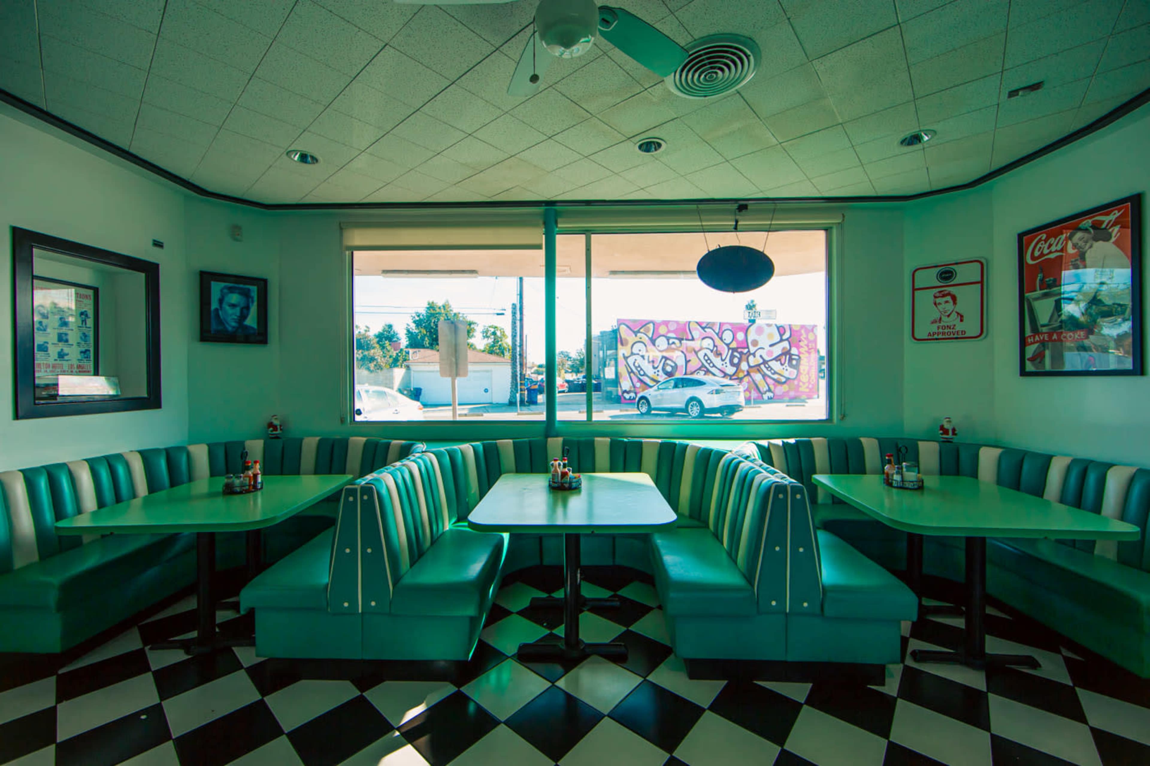 The image shows a diner with teal booths and black-and-white checkered flooring, featuring large windows that overlook a mural on the opposite wall.