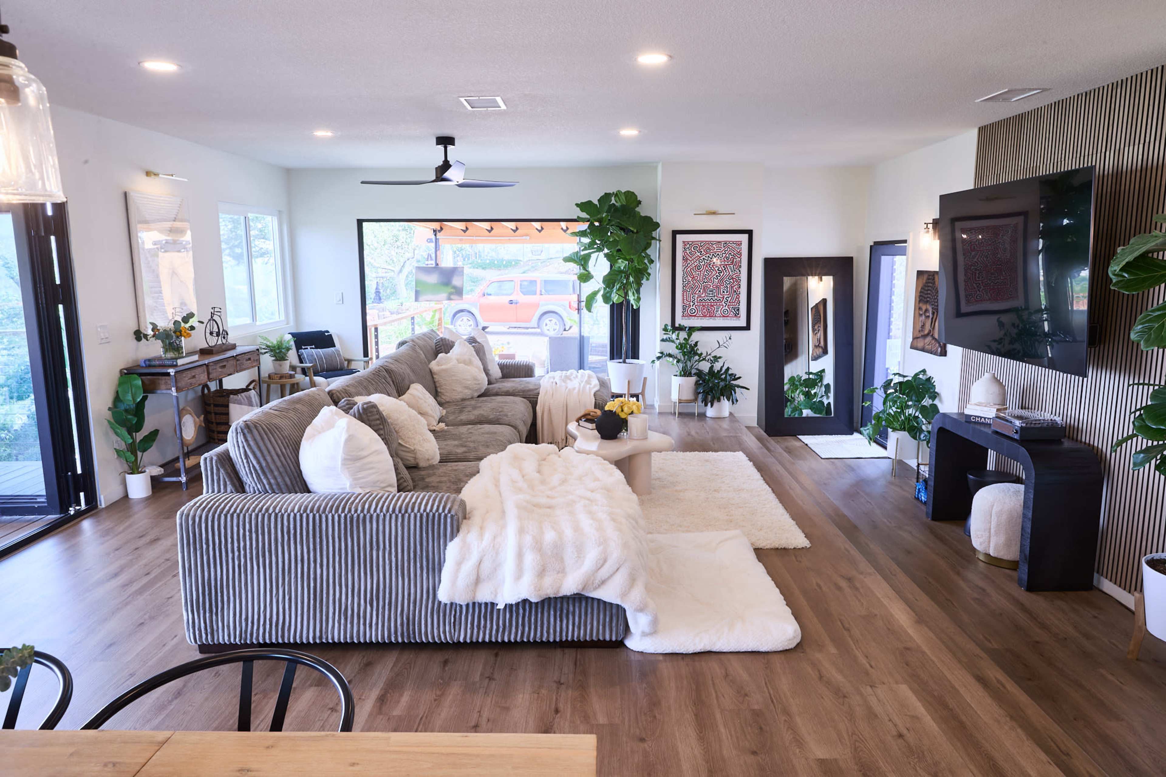 The living room features a large gray sectional sofa with white pillows, a plush rug, and a television mounted on the wall, surrounded by indoor plants and natural light from large windows.