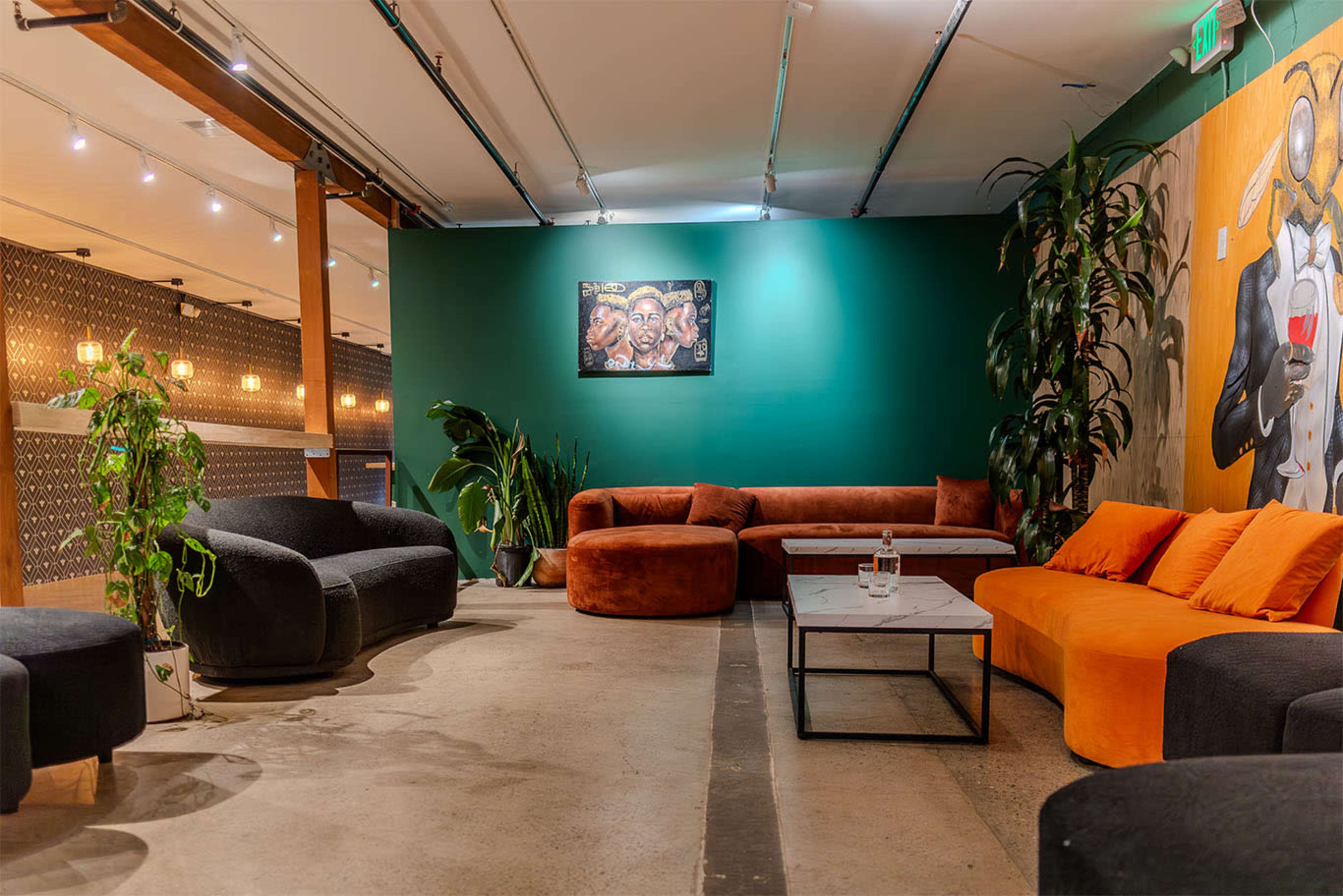 The image shows a stylish lounge area with black and orange sofas, a coffee table, plants, and a green wall featuring a piece of art.